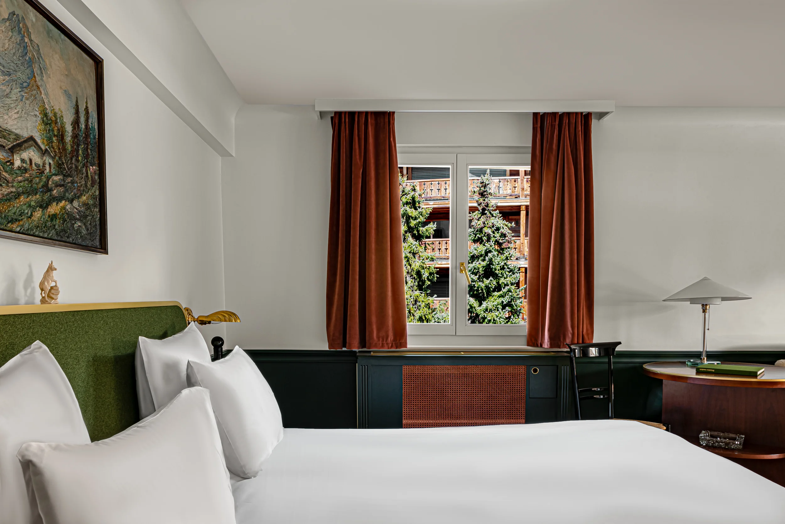 Camera d'albergo con letto bianco, testiera verde, pittura, tende marroni e finestra con vista sui pini.