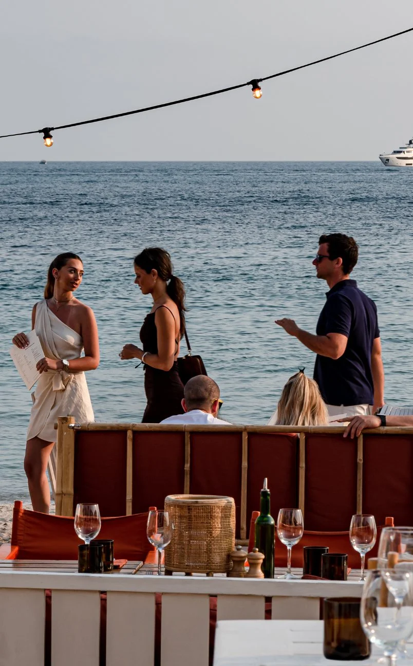 La gente socializa en la playa con mesas y luces de cadena encendidas al atardecer.