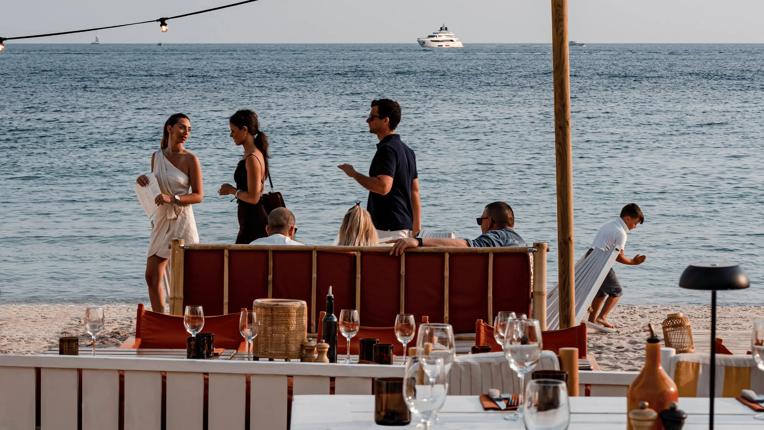 Gente socializando y relajándose en un restaurante junto a la playa con el océano y los barcos de fondo.