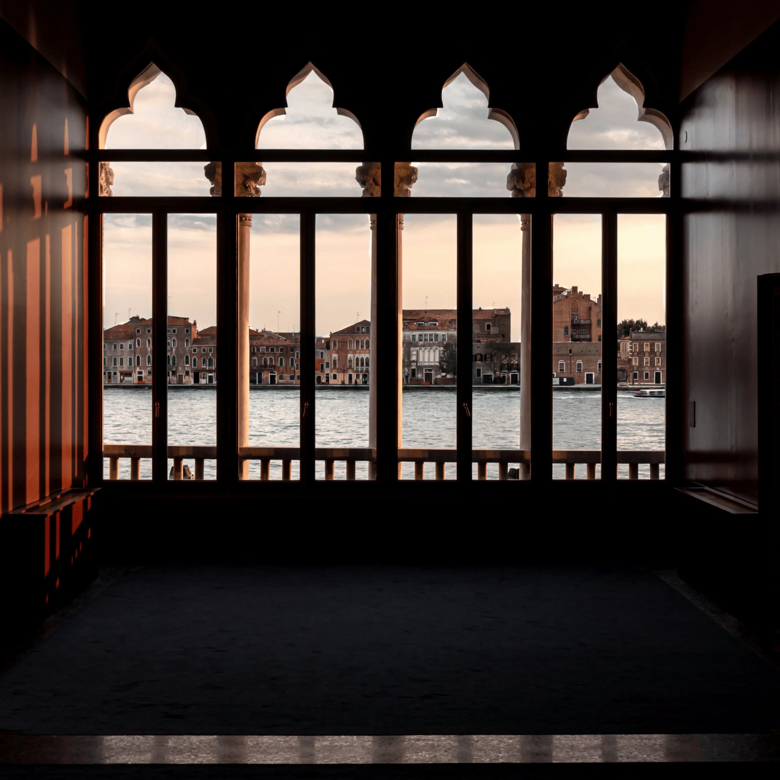 View of Venetian buildings across a canal through arched windows of a dark interior at sunset.