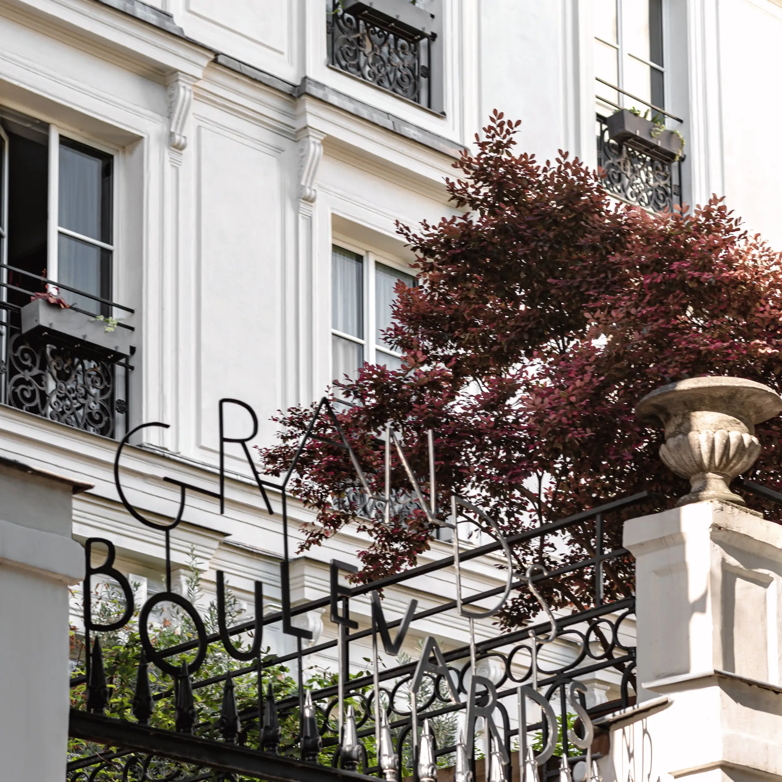 Ornate black metal gate with GRANDS BOULEVARDS text in front of a white building and red-leaf tree.