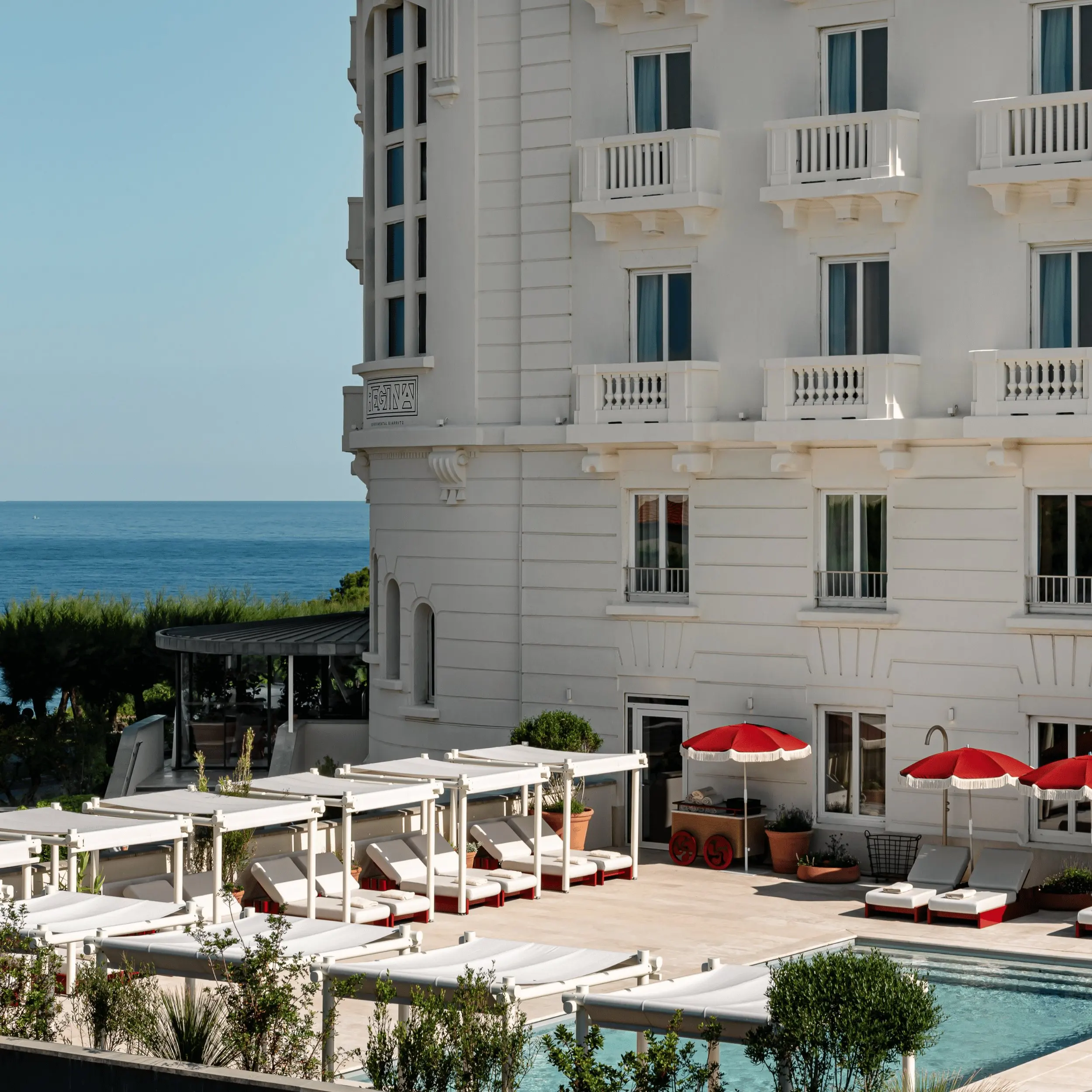 White hotel building by pool with lounge chairs, red umbrellas, and ocean view in the background.