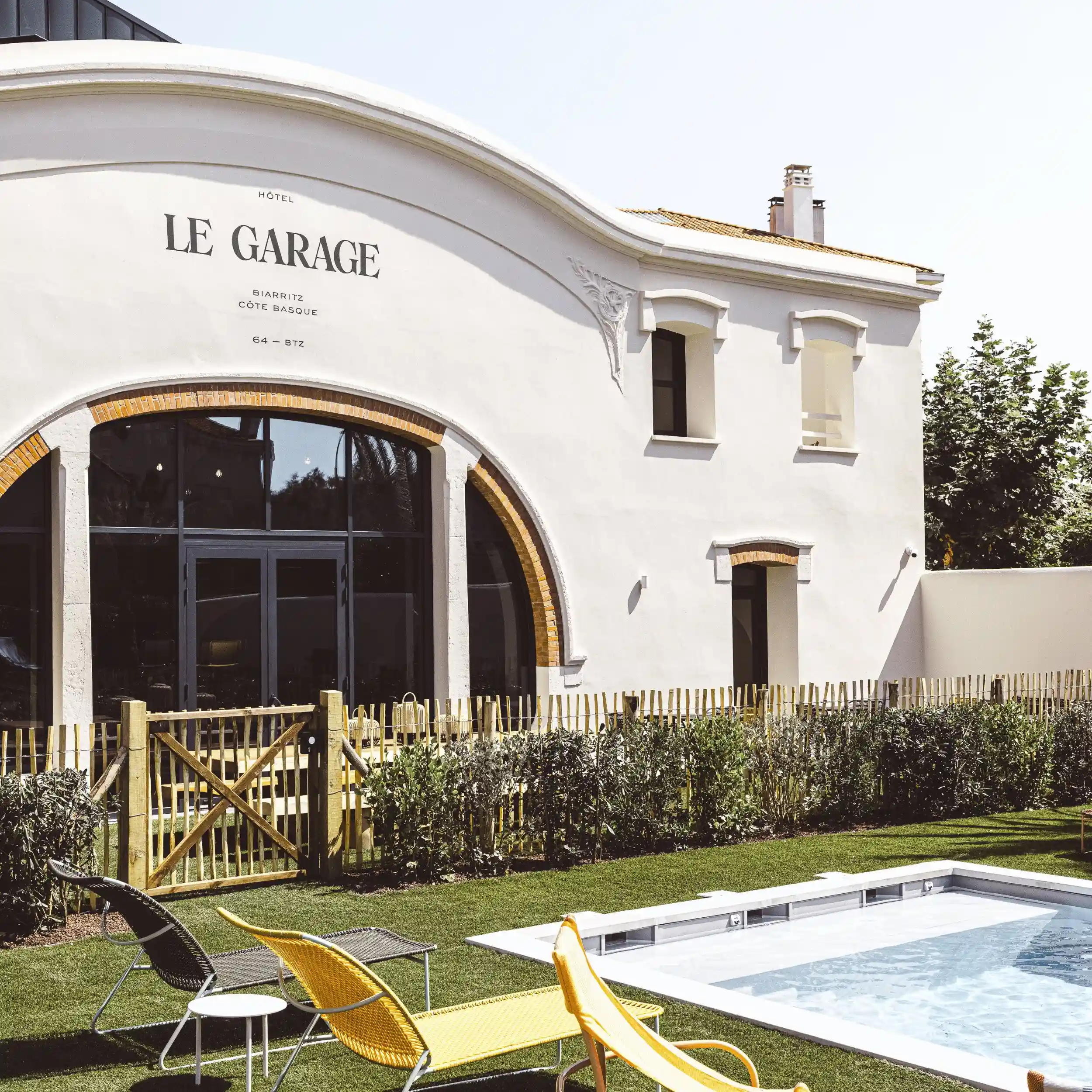 White building with large arched window, garden, yellow lounge chairs, and a small pool.