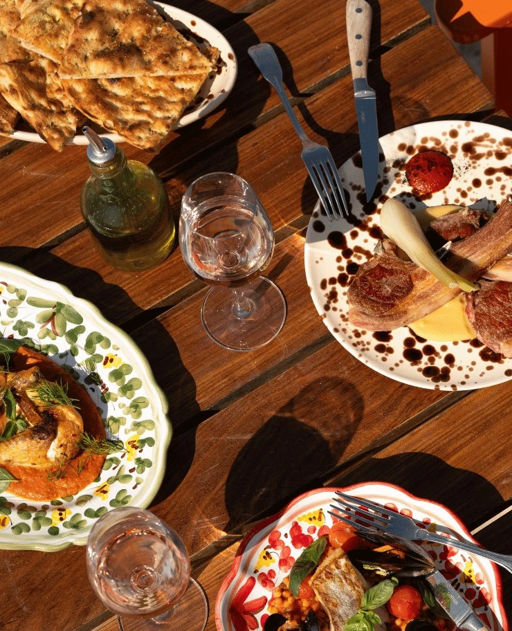 Mesa con platos de carne, pescado, pan a la parrilla, botella de aceite de oliva y dos copas de vino sobre superficie de madera.
