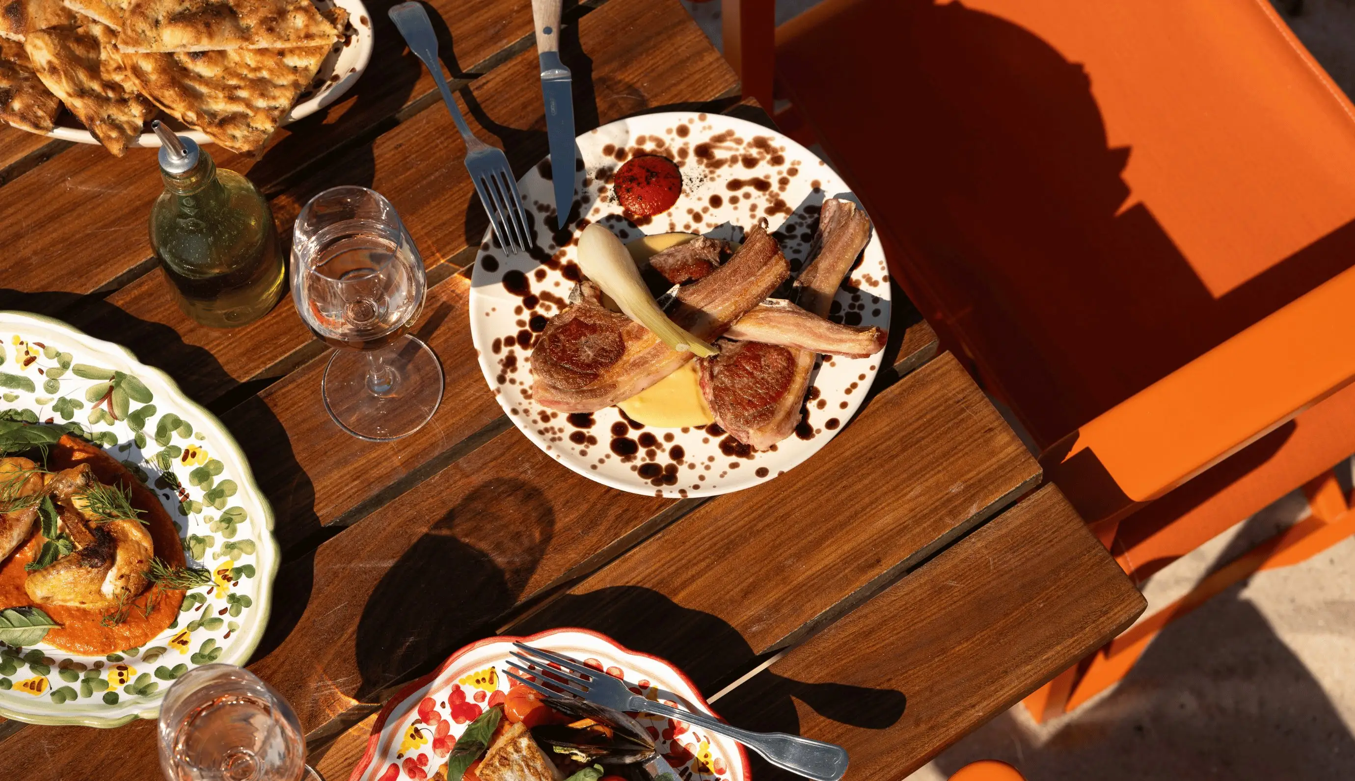 Mesa con tres platos de comida gourmet, dos vasos de agua, una botella de aceite de oliva y una silla naranja cerca.