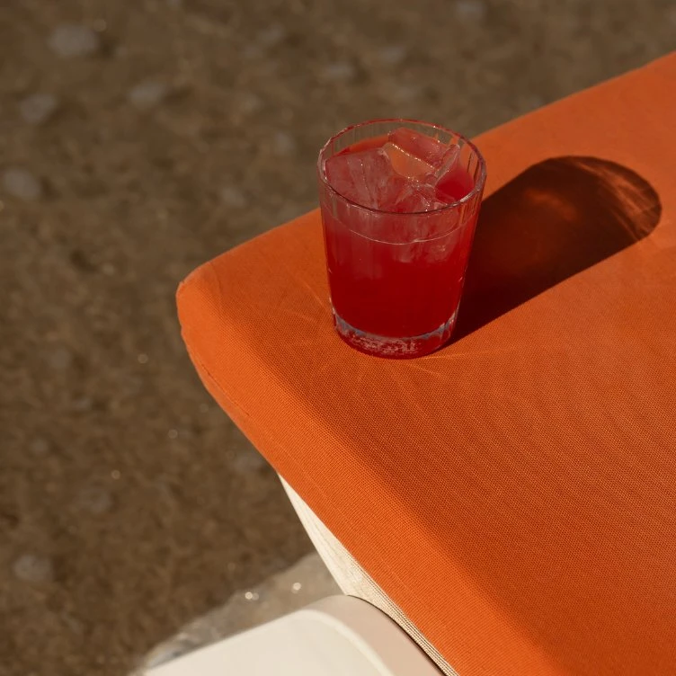 Bebida roja con cubitos de hielo en un vaso sobre una superficie de tela naranja que proyecta una sombra.