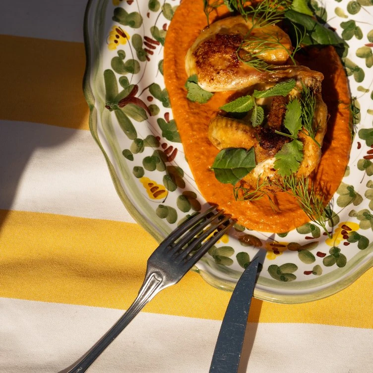 Pollo asado en salsa roja adornado con hierbas, servido en un plato floral con tenedor y cuchillo.