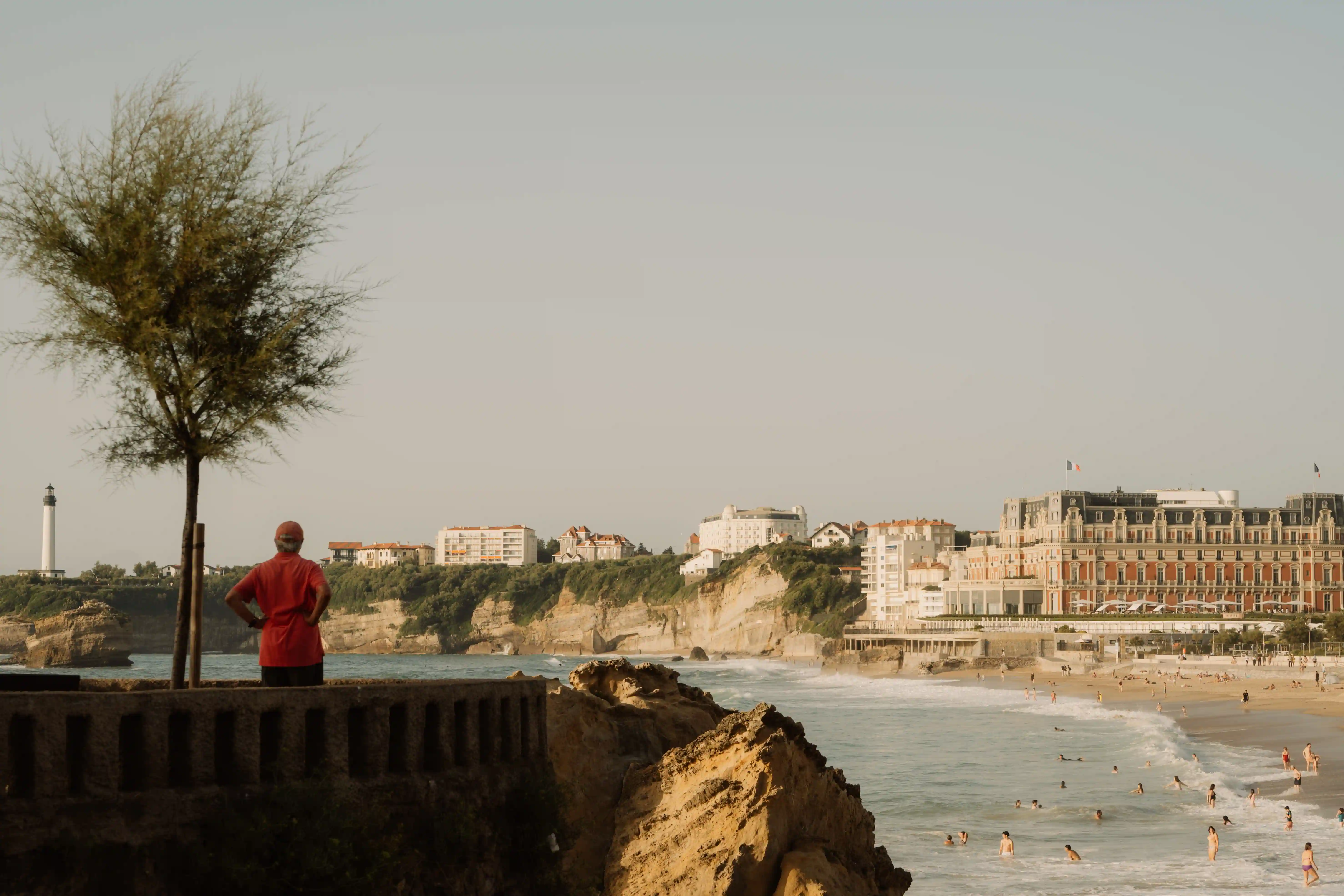 Persona in camicia rossa in piedi vicino ad un albero con vista sulla spiaggia con nuotatori e edifici sul mare.