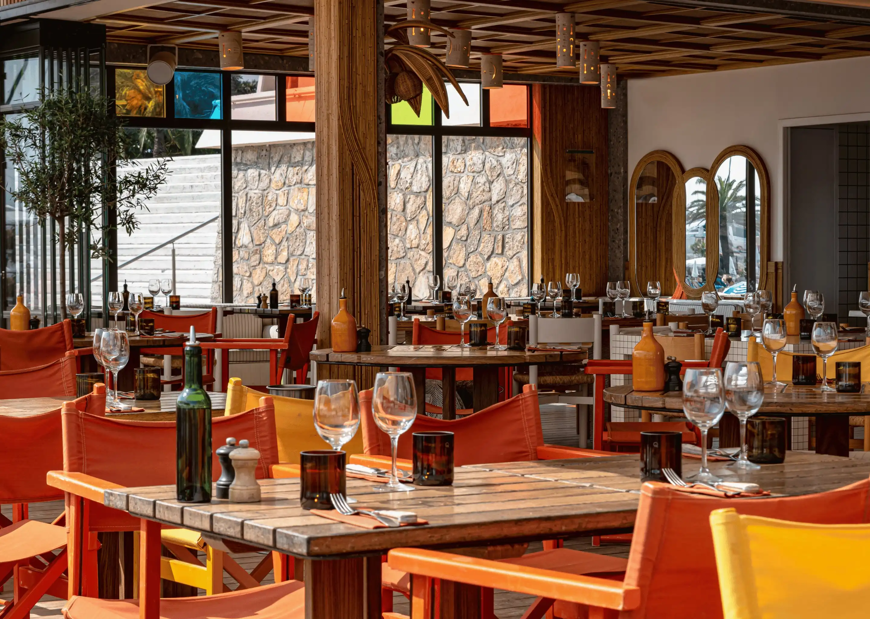 Área de comedor vacía del restaurante con mesas de madera, sillas coloridas, cristalería y ventanas de piedra.
