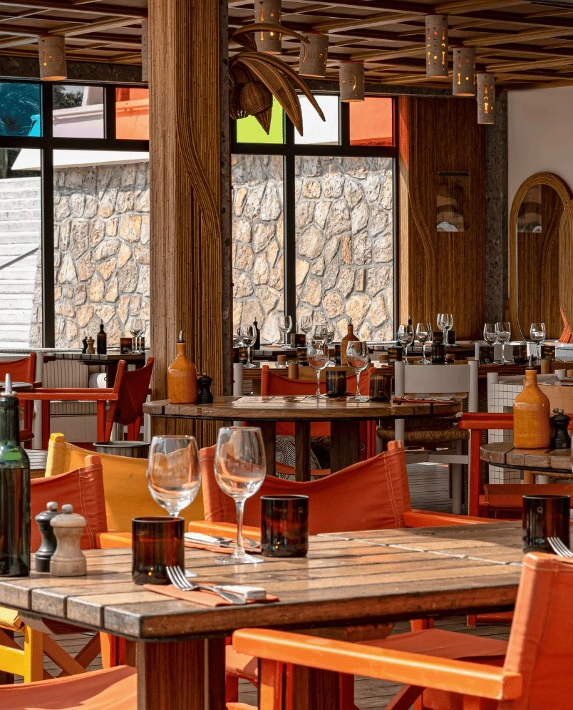 Restaurante interior con mesas de madera, sillas naranjas, cristalería y pared de piedra visible a través de las ventanas.