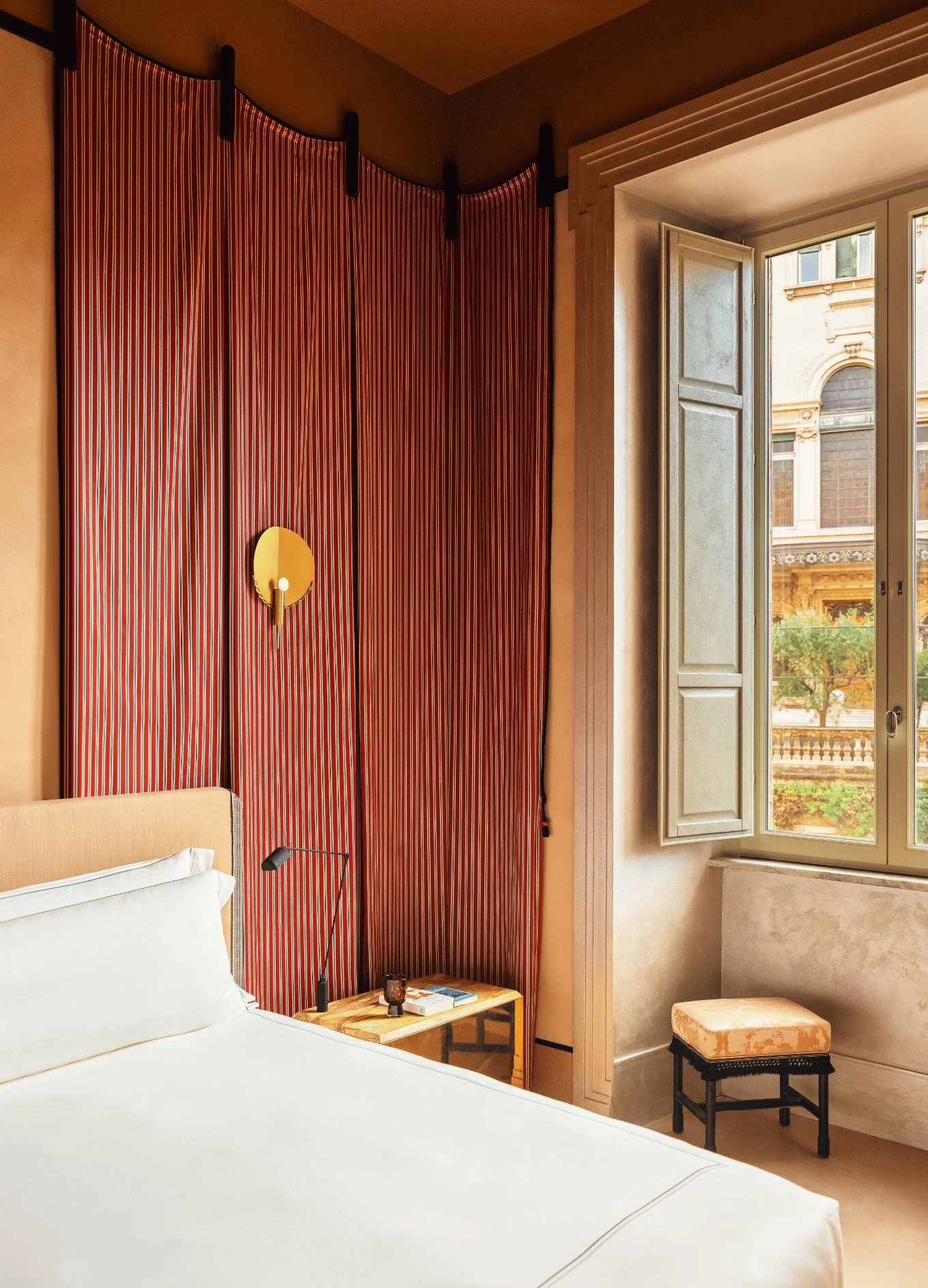 Angolo della camera da letto con letto bianco, tende a righe rosse, applique dorate e finestra aperta con vista sull'architettura.