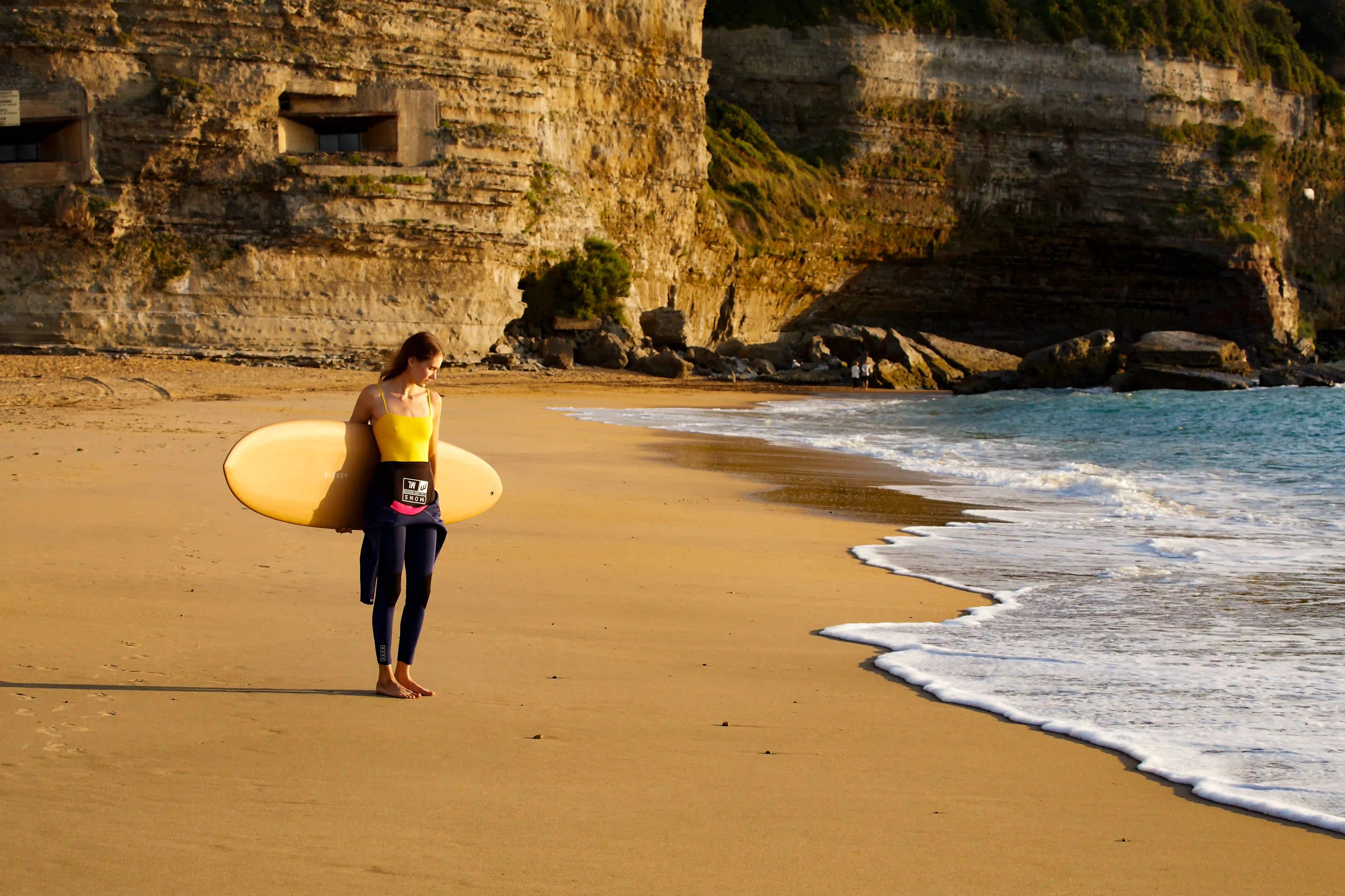 Donna con un top giallo che tiene una tavola da surf che cammina a piedi nudi su una spiaggia sabbiosa vicino a scogliere e oceano.