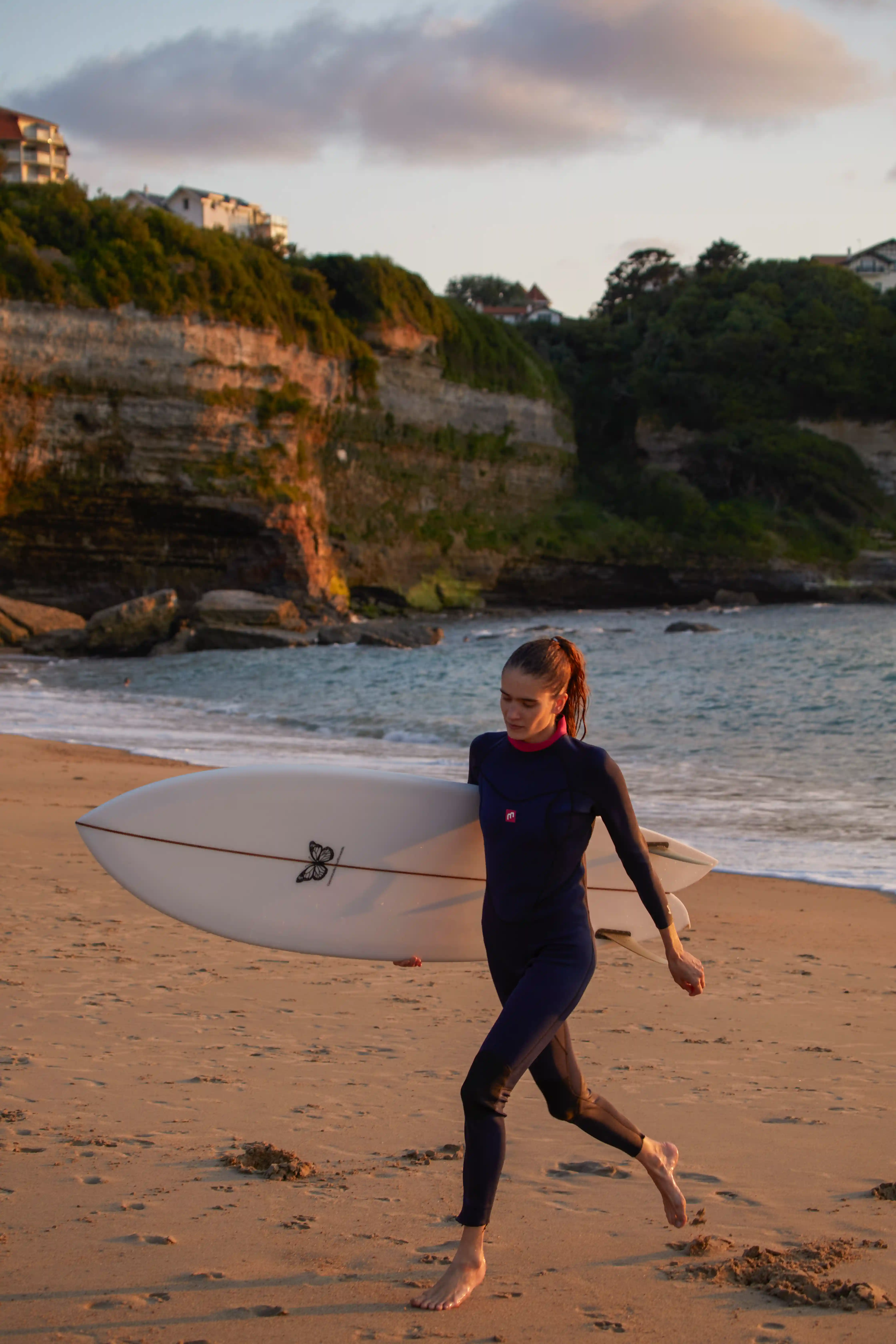 Donna in muta che trasporta una tavola da surf che cammina a piedi nudi su una spiaggia sabbiosa vicino alle scogliere al tramonto.