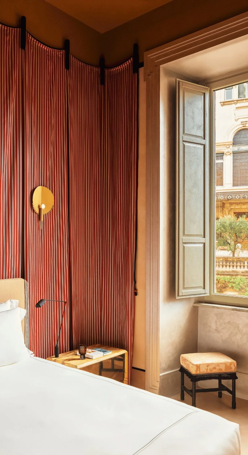 Angolo della camera d'albergo con tenda a righe rosse, letto bianco, applique dorata e finestra aperta con sgabello esterno.