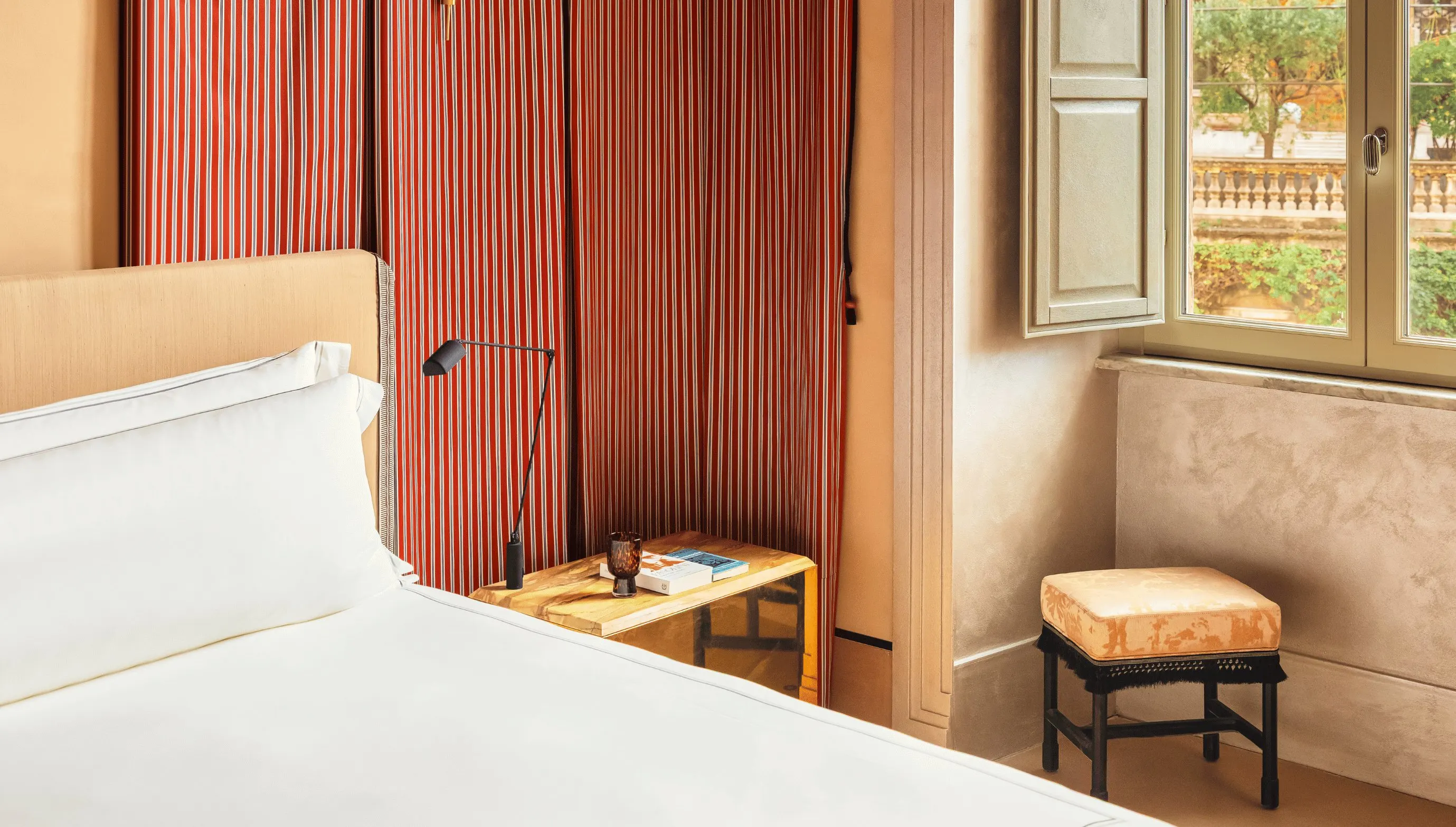 Angolo della camera d'albergo con letto bianco, tavolino in legno, lampada, tenda rossa a righe, finestra aperta e sgabello.