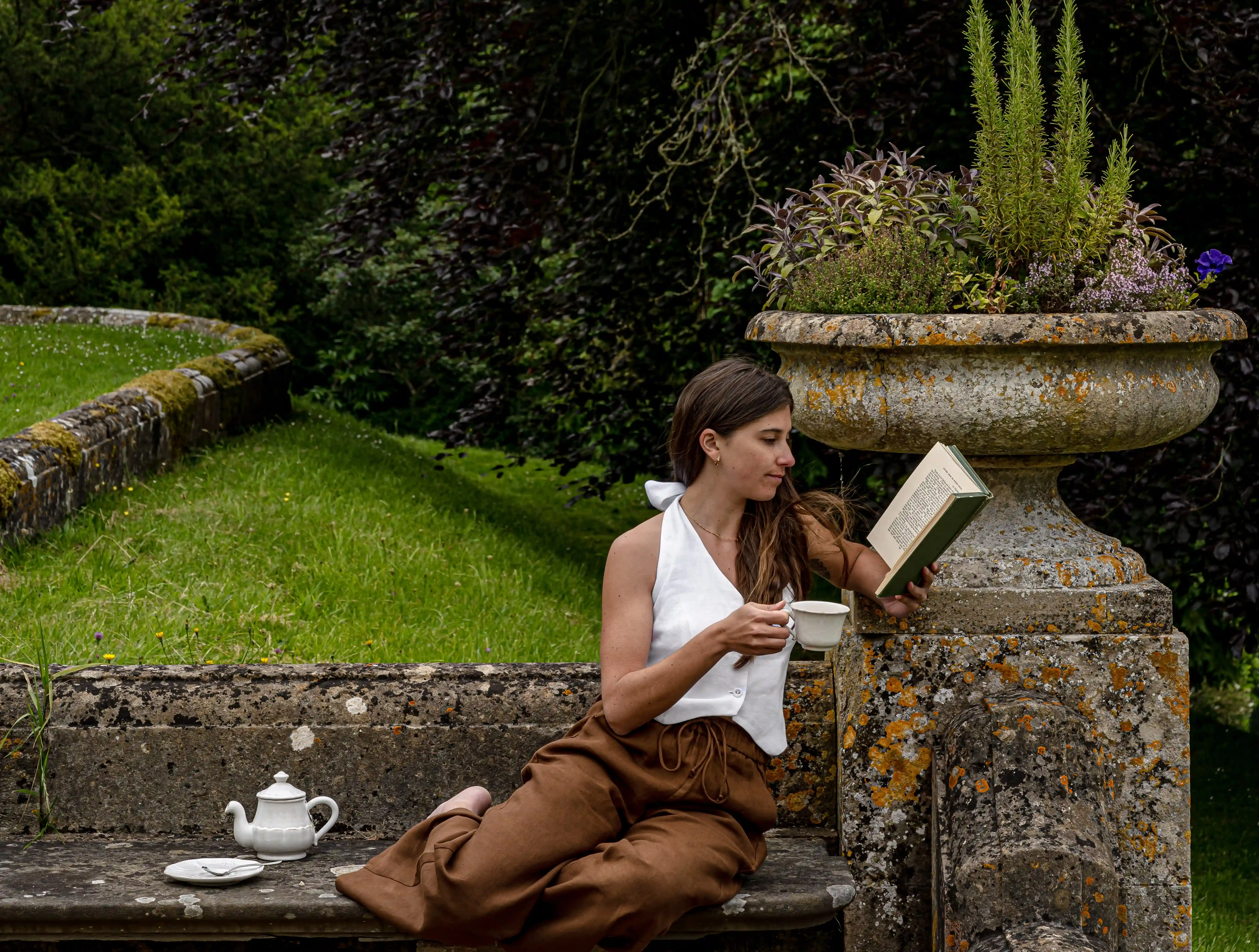 La donna in top bianco e pantaloni marroni legge un libro e tiene una tazza da tè seduta su una panca di pietra.