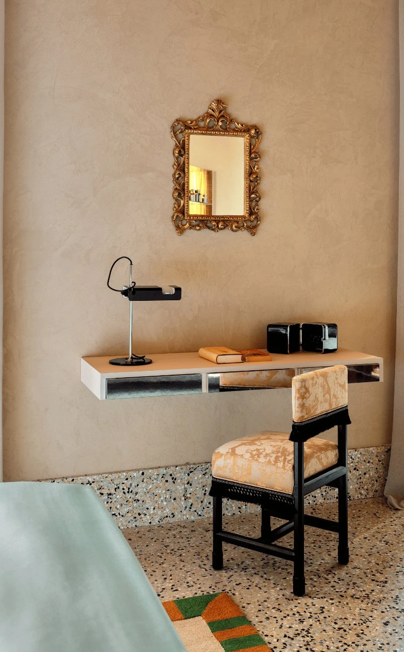 Espacio de trabajo minimalista con pared de color beige, espejo adornado, escritorio flotante, silla vintage y una lámpara pequeña.