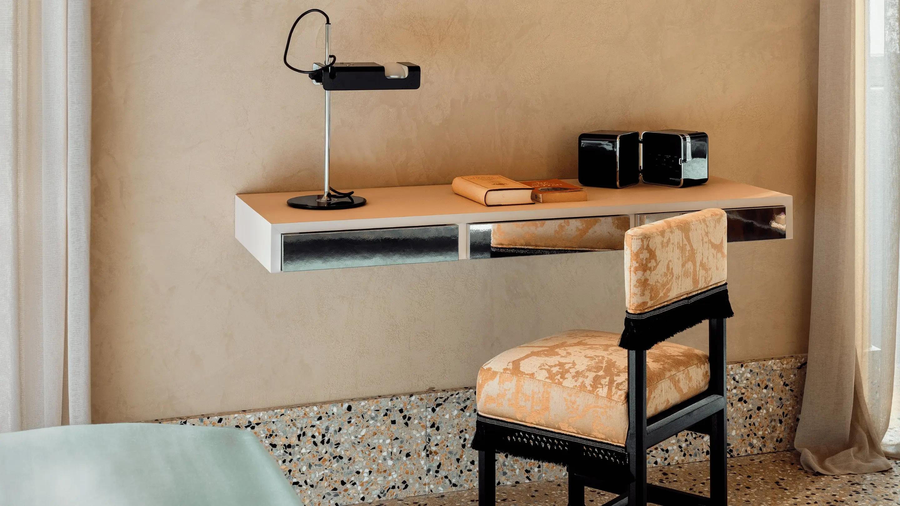 Escritorio minimalista de color beige con cajones con espejo, una lámpara negra, libros y una silla tapizada con flecos.