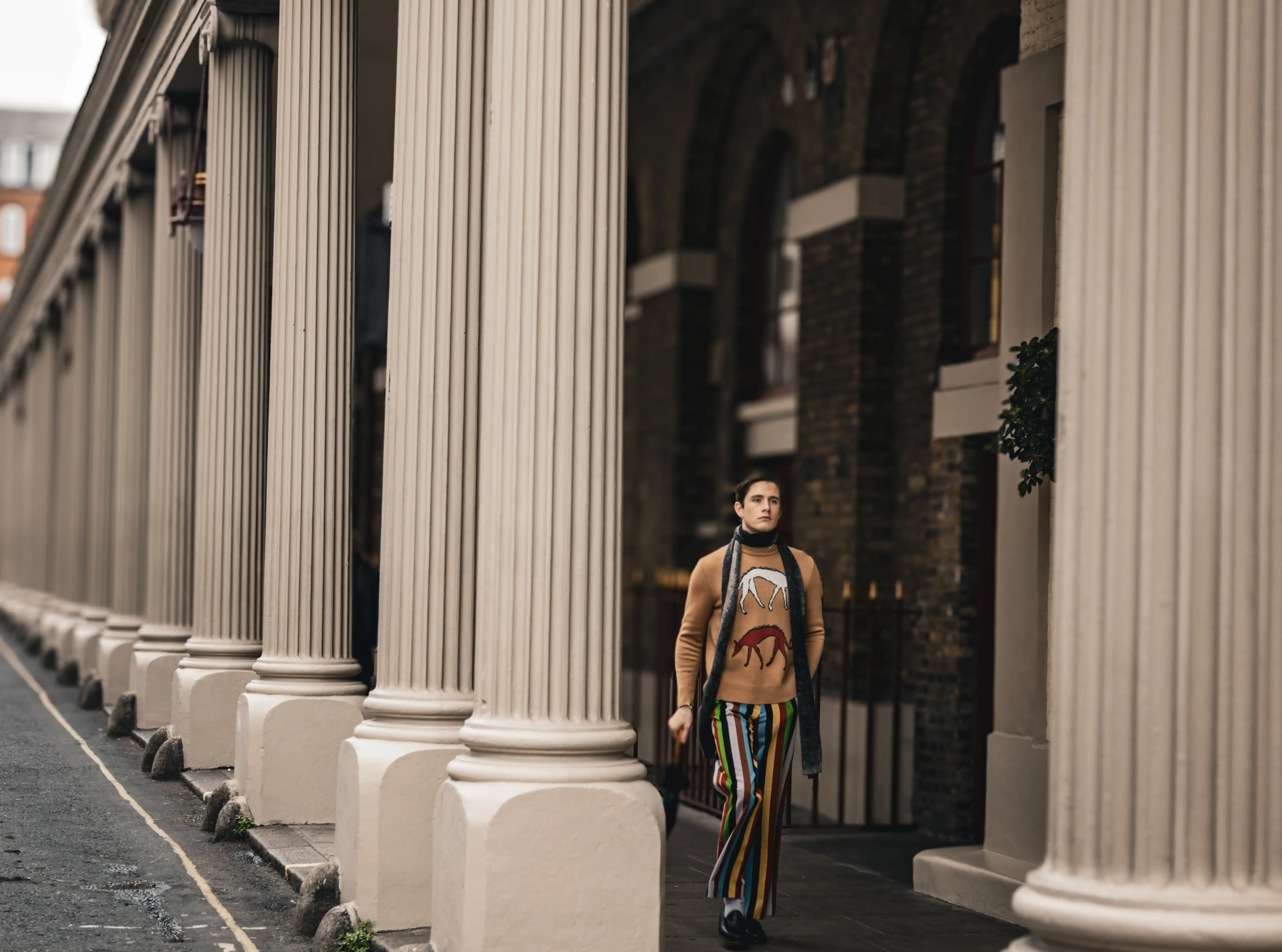 Persona que camina junto a altas columnas de color beige con pantalones a rayas y un suéter marrón con diseños de animales.