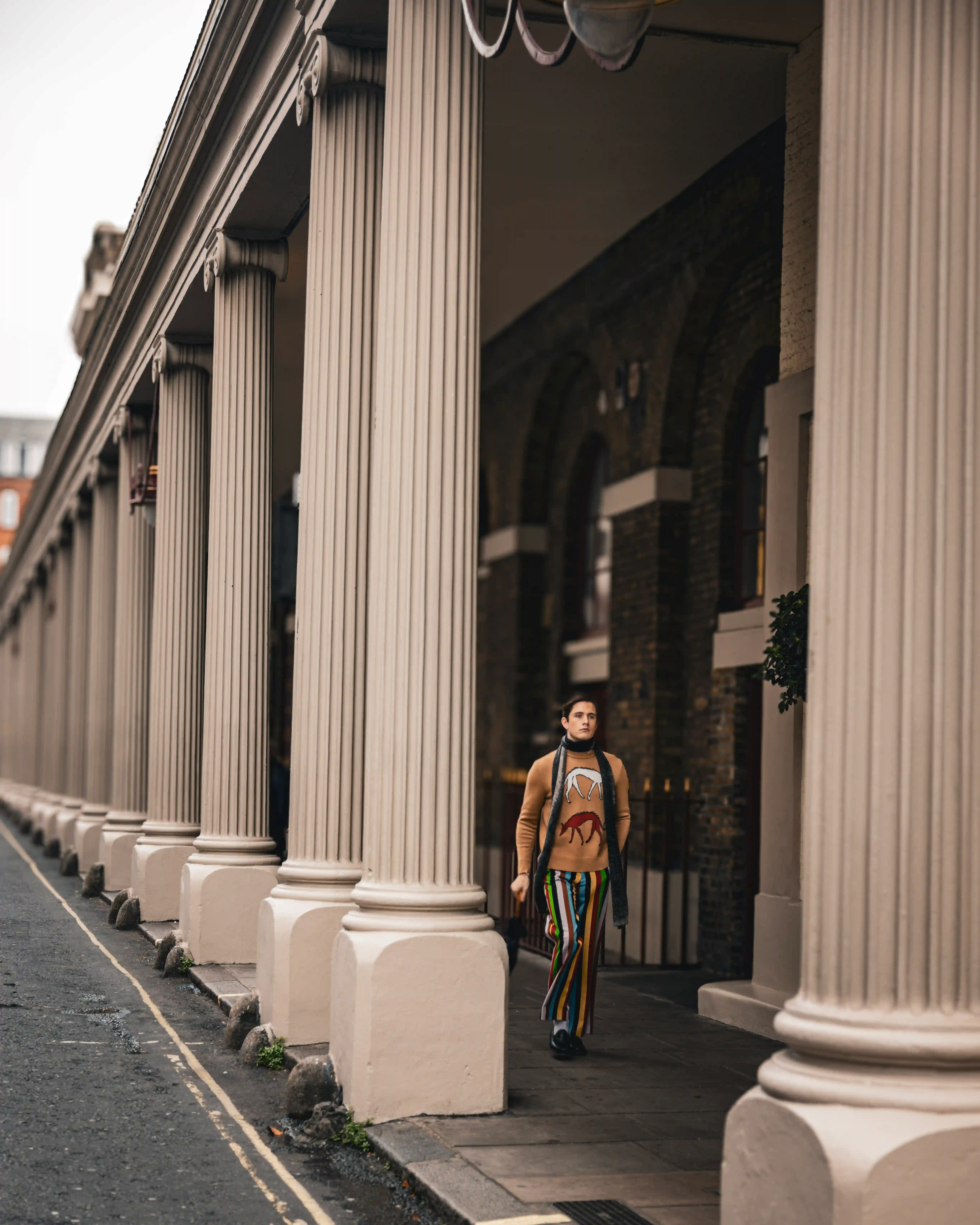 Persona con pantalones a rayas y suéter con estampado animal que camina junto a las grandes columnas de un edificio clásico.