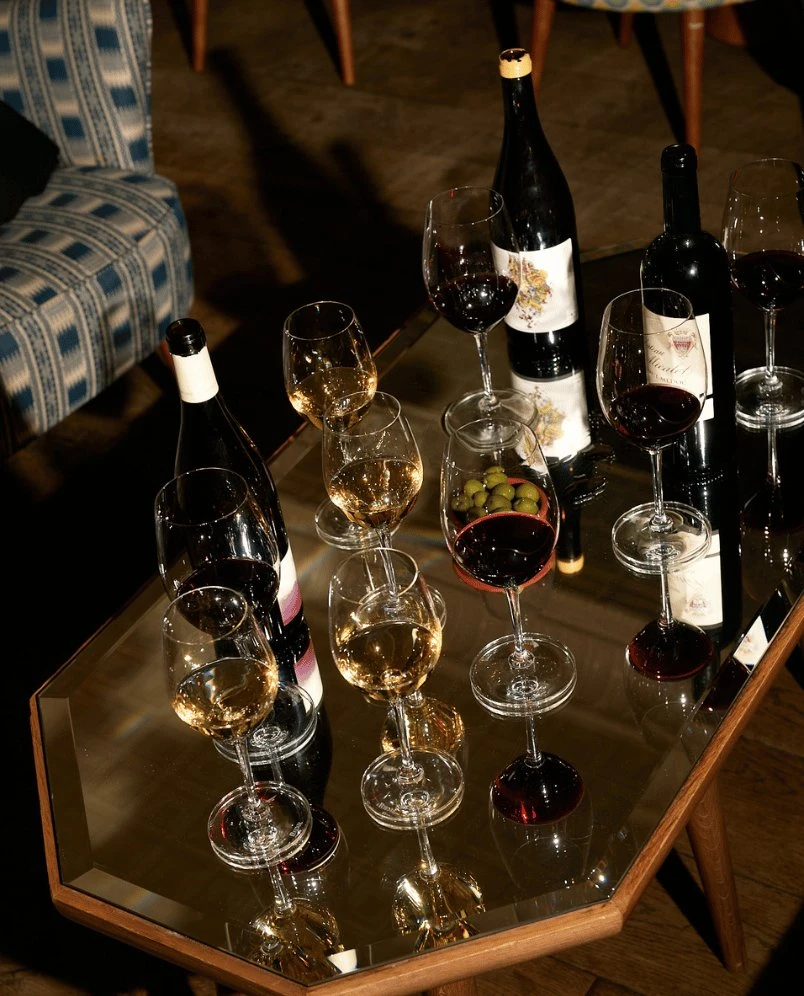 Mesa com várias taças de vinho tinto e branco, garrafas de vinho e uma tigela de vidro com azeitonas.