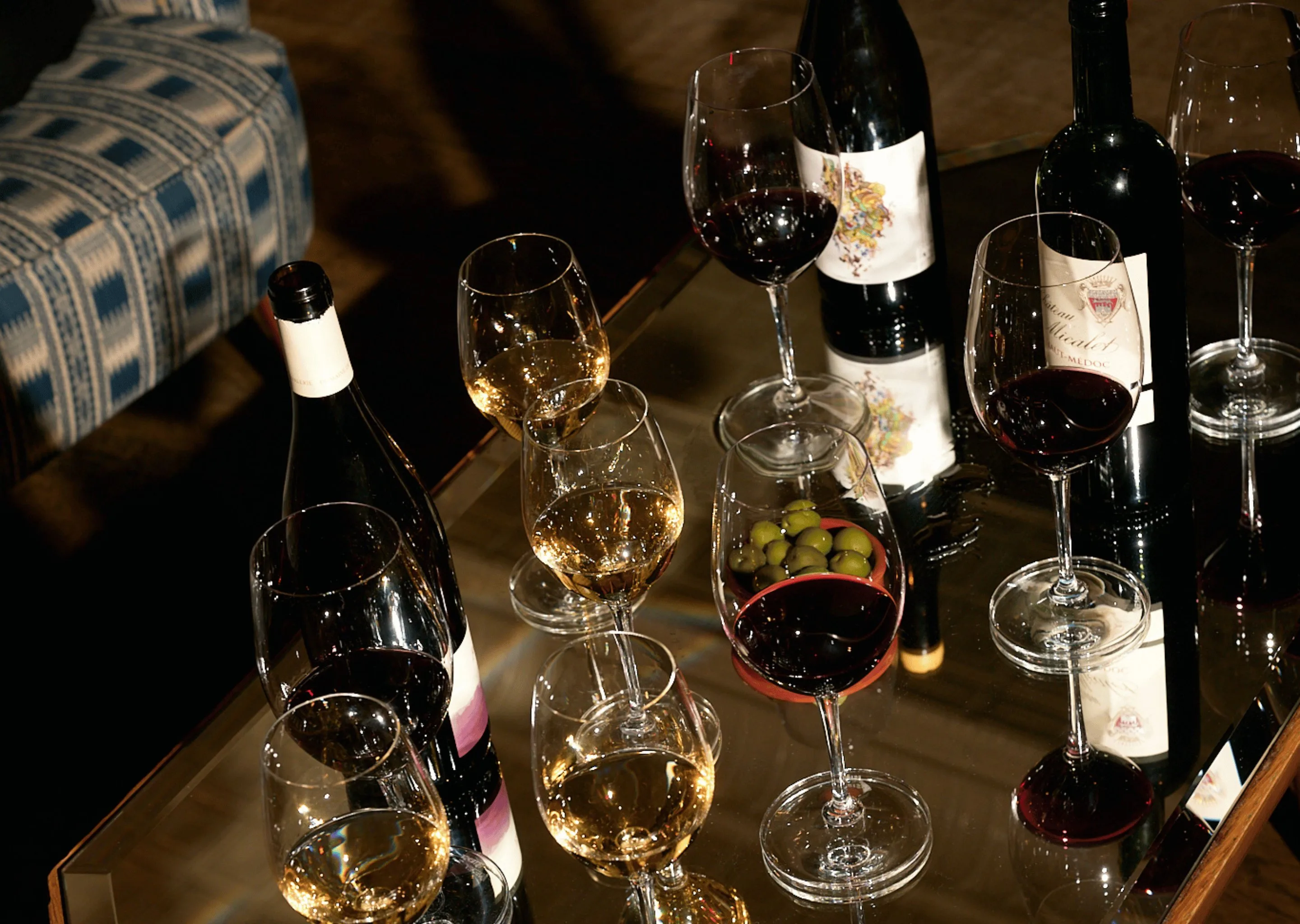 Mesa com taças de vinho tinto e branco, garrafas de vinho e uma tigela de azeitonas verdes.