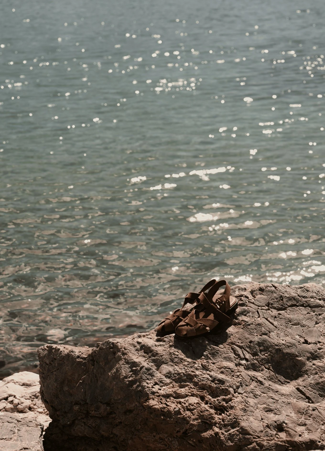 Sandalias tejidas marrones colocadas sobre una roca junto al agua cristalina del mar en la playa.