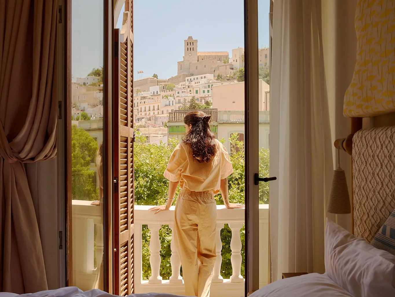 La donna in abito beige si trova sul balcone guardando la città collinare con edifici storici.