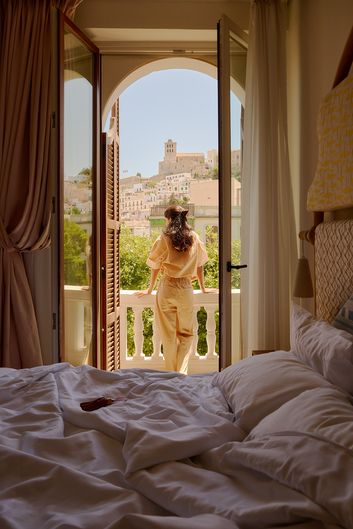 La persona in abito beige si trova sul balcone guardando una città collinare sotto un cielo limpido.