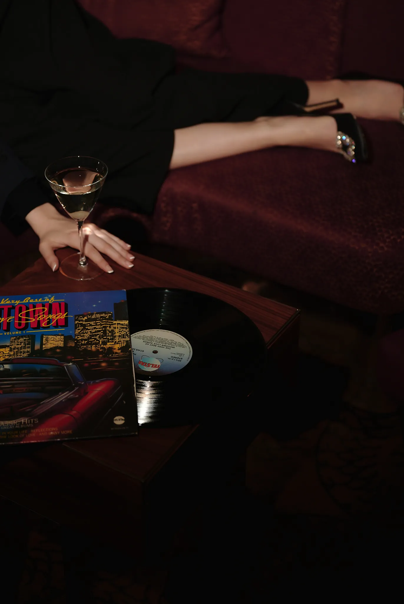 Person reclining on red couch with martini glass, vinyl record and album cover on wooden table.