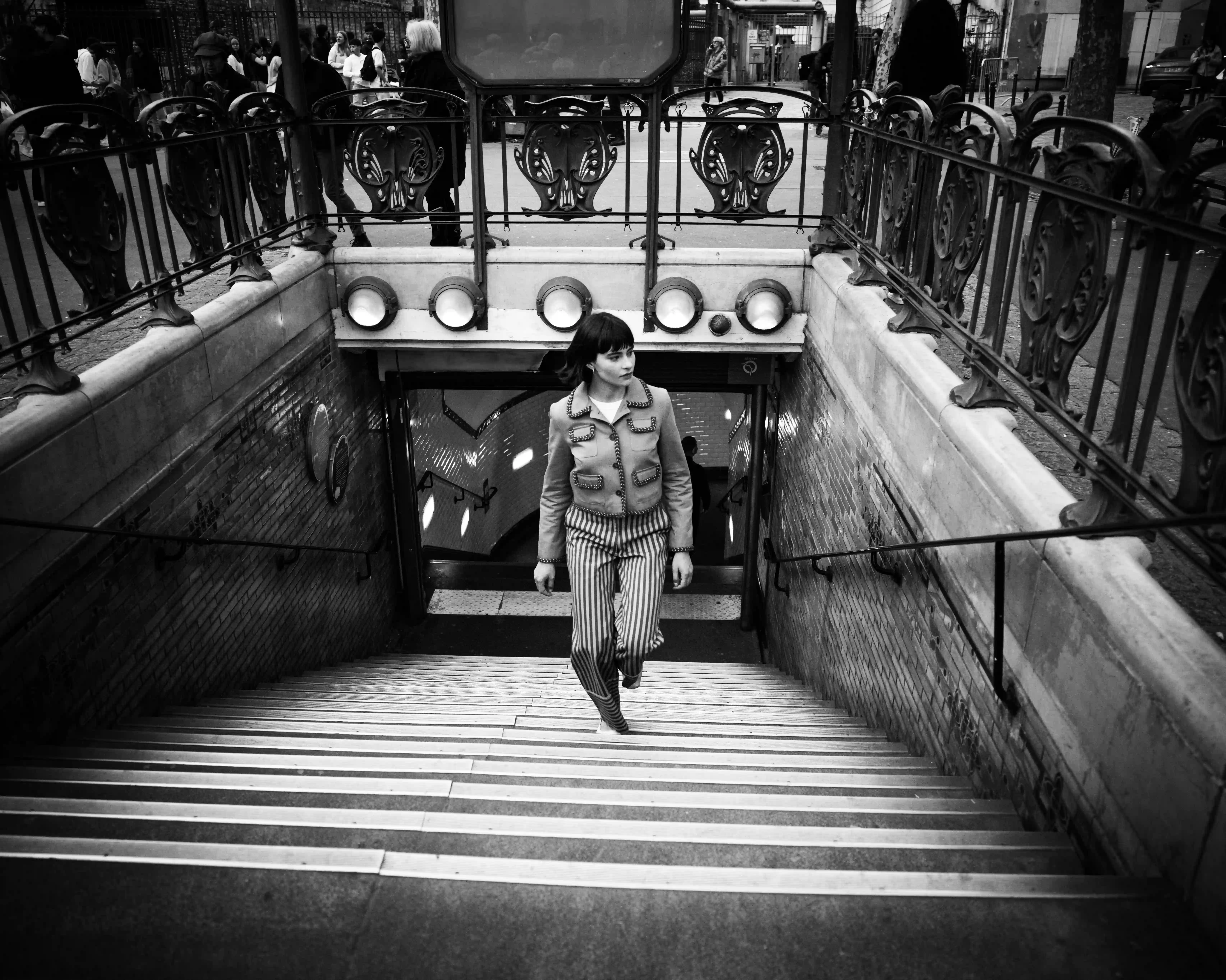 Mujer con pantalones a rayas que sube las escaleras desde la entrada de un metro con barandillas de hierro adornadas en blanco y negro.