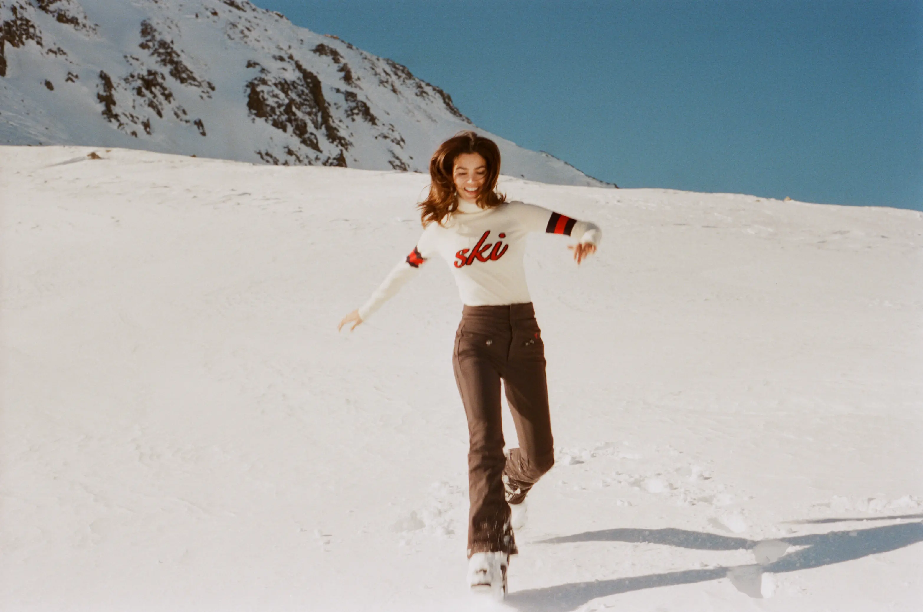Mulher caminhando alegremente na neve em roupa de esqui com fundo de montanha sob céu azul claro.