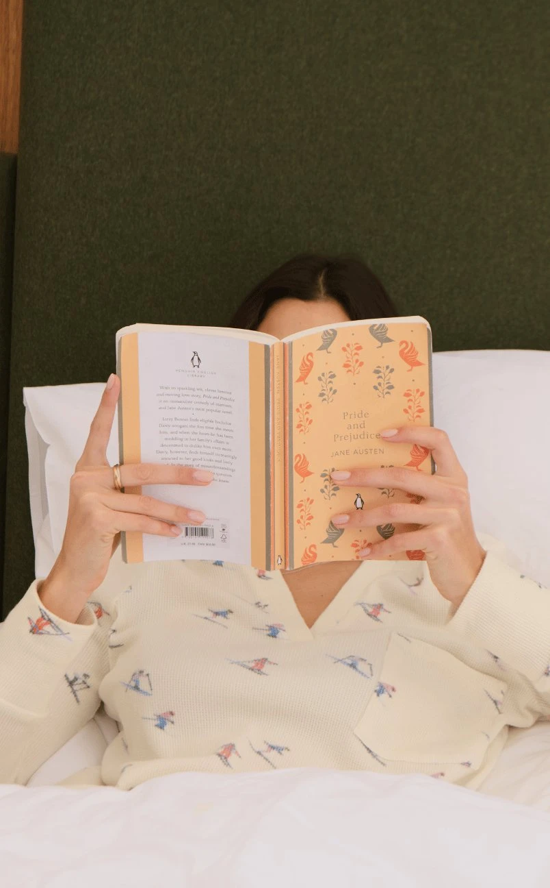 Pessoa deitada na cama lendo uma cópia de Orgulho e Preconceito de Jane Austen com uma capa estampada.