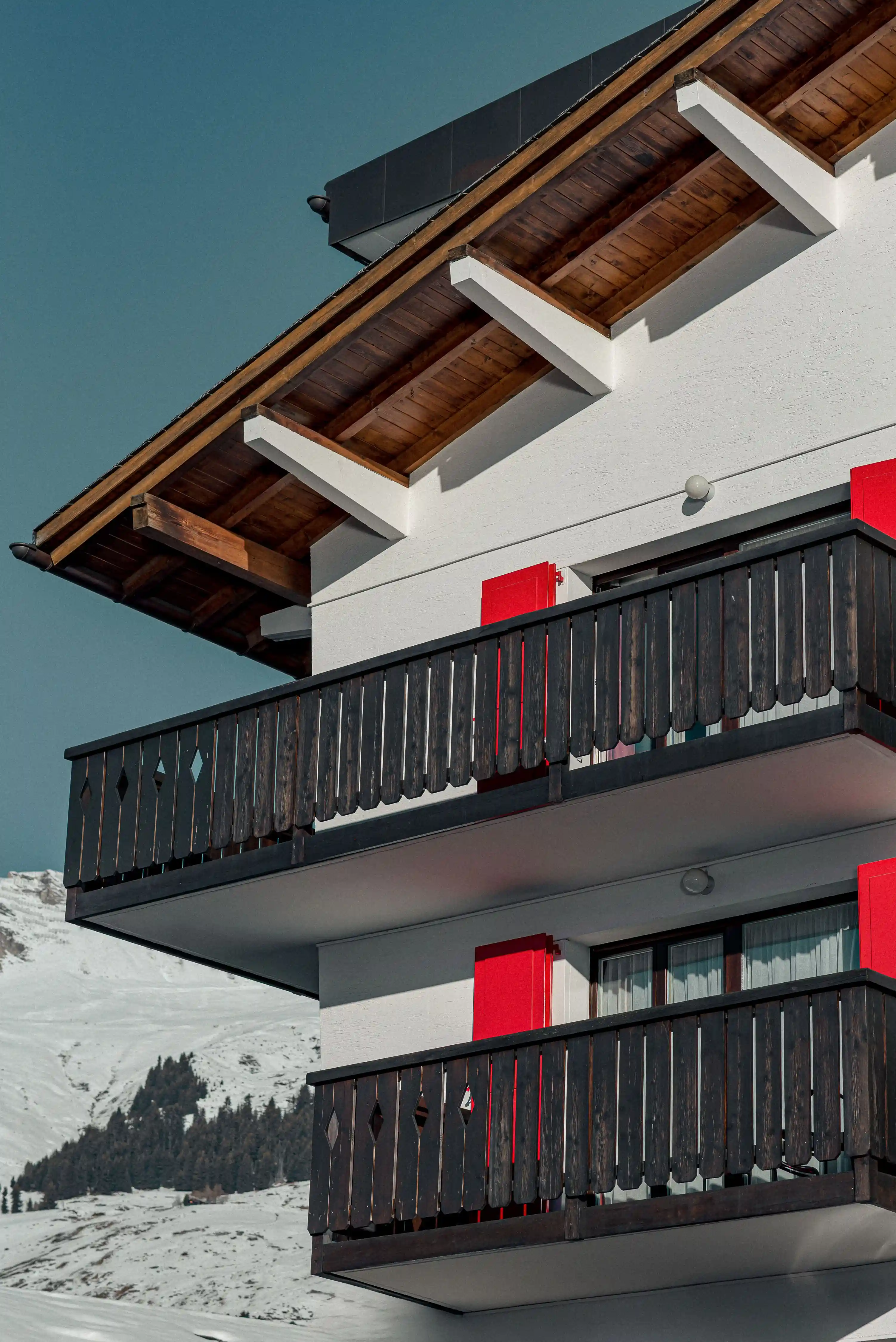 Close-up view of a chalet balcony with red shutters and snowy mountain background.