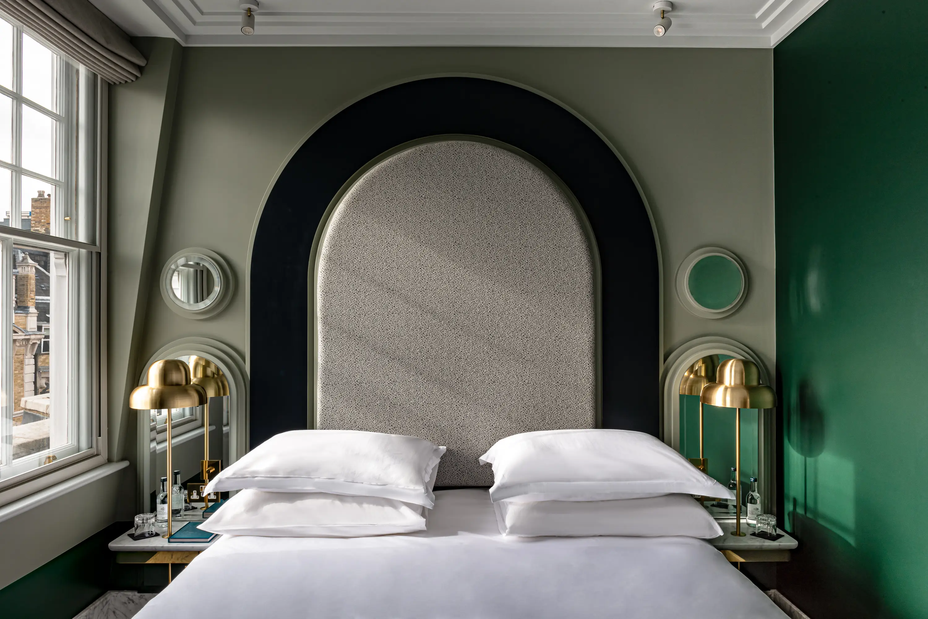 Modern hotel bedroom with large arched headboard, two gold lamps, and a window showing city buildings.