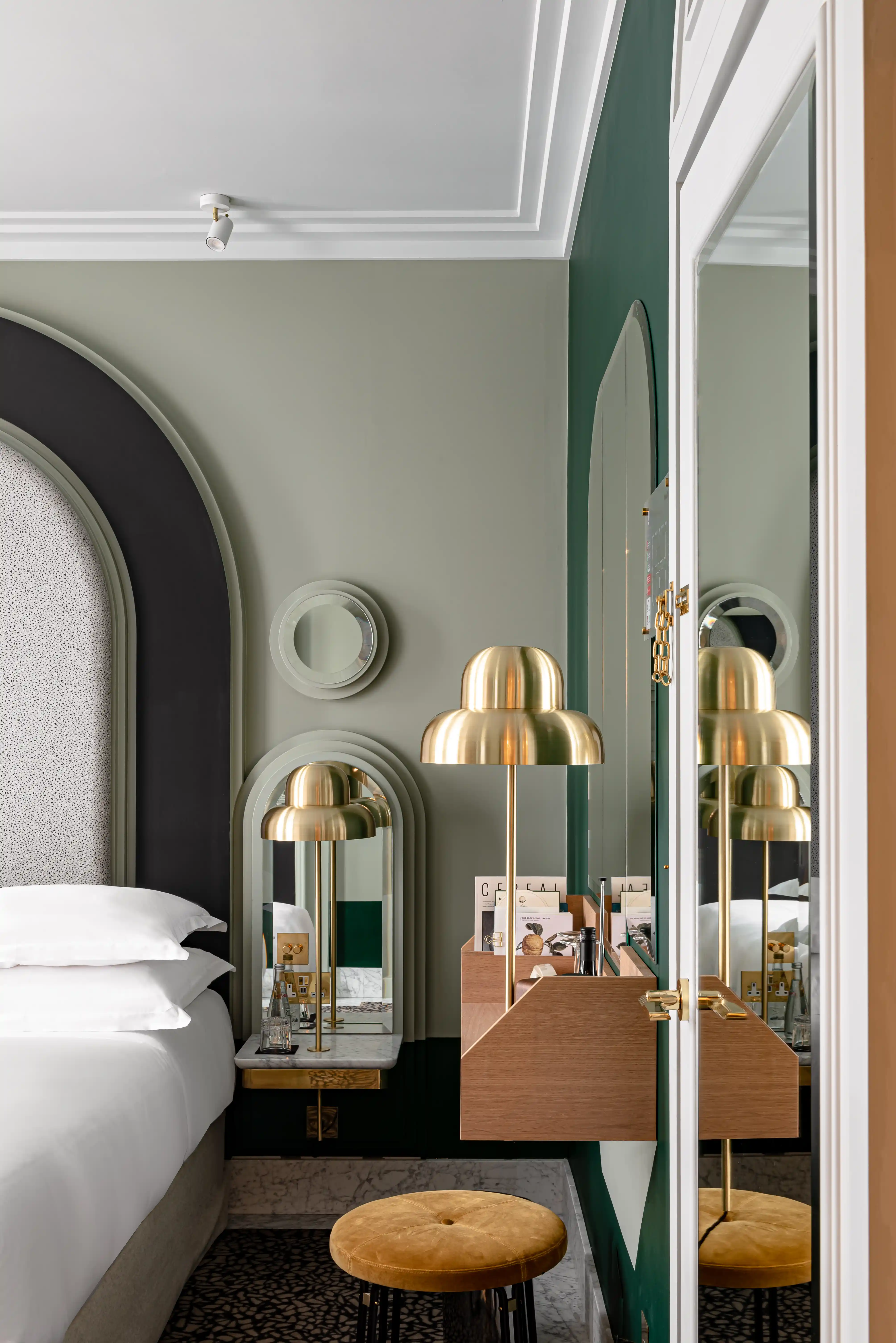 Hotel bedroom corner with white bed, gold lamp, round stool, mirrors, and green walls.