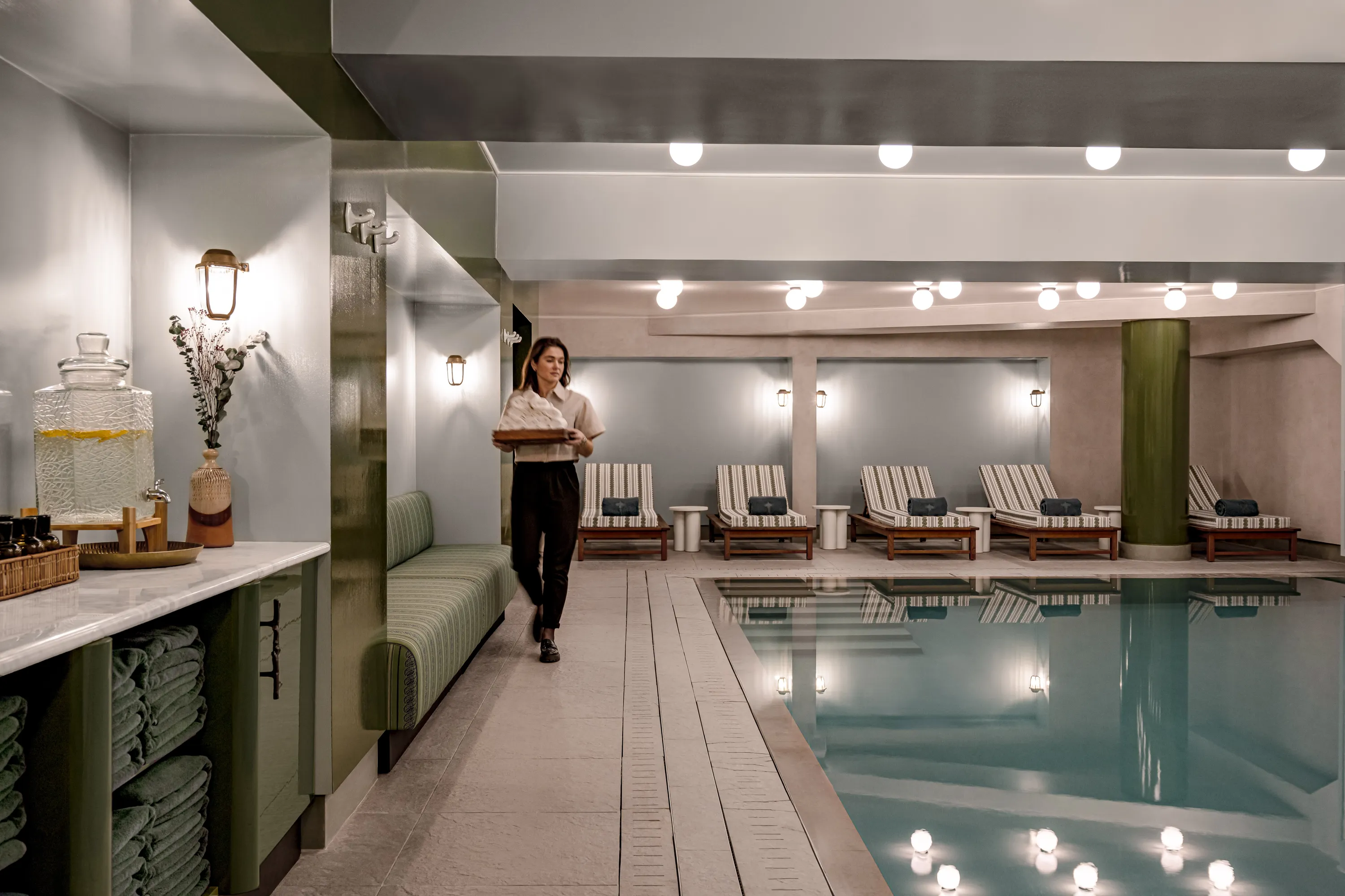 Woman walking by an indoor pool with striped lounge chairs and green towel shelves.