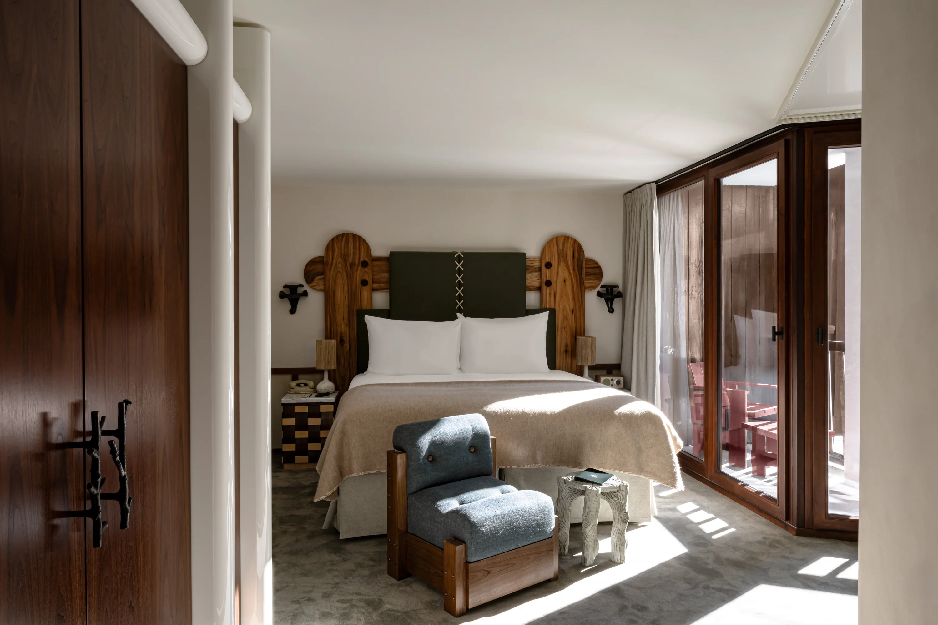 Hotel bedroom with large bed, wooden headboard, blue chair, and glass doors opening to balcony.