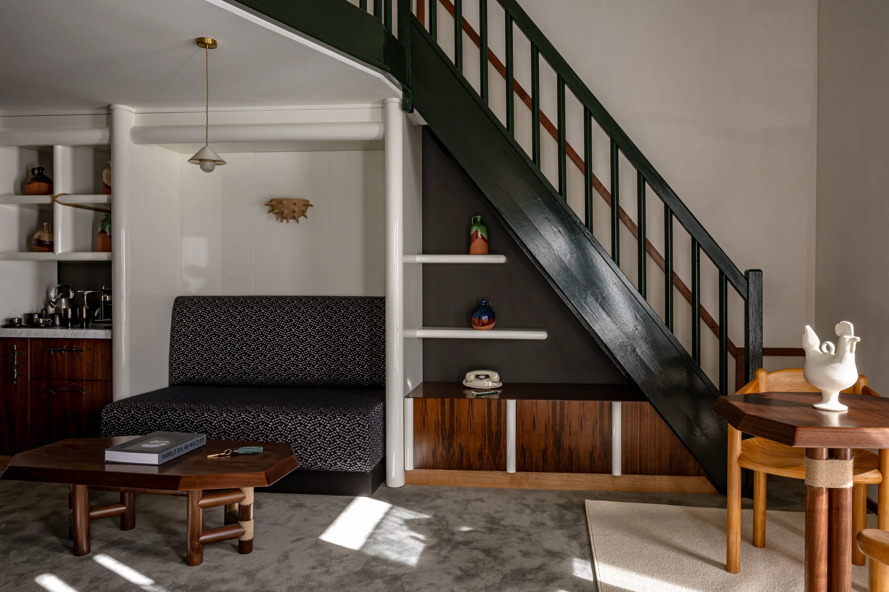 Cozy seating area with patterned sofa, wooden table, dark staircase, shelves, and decorative pottery.