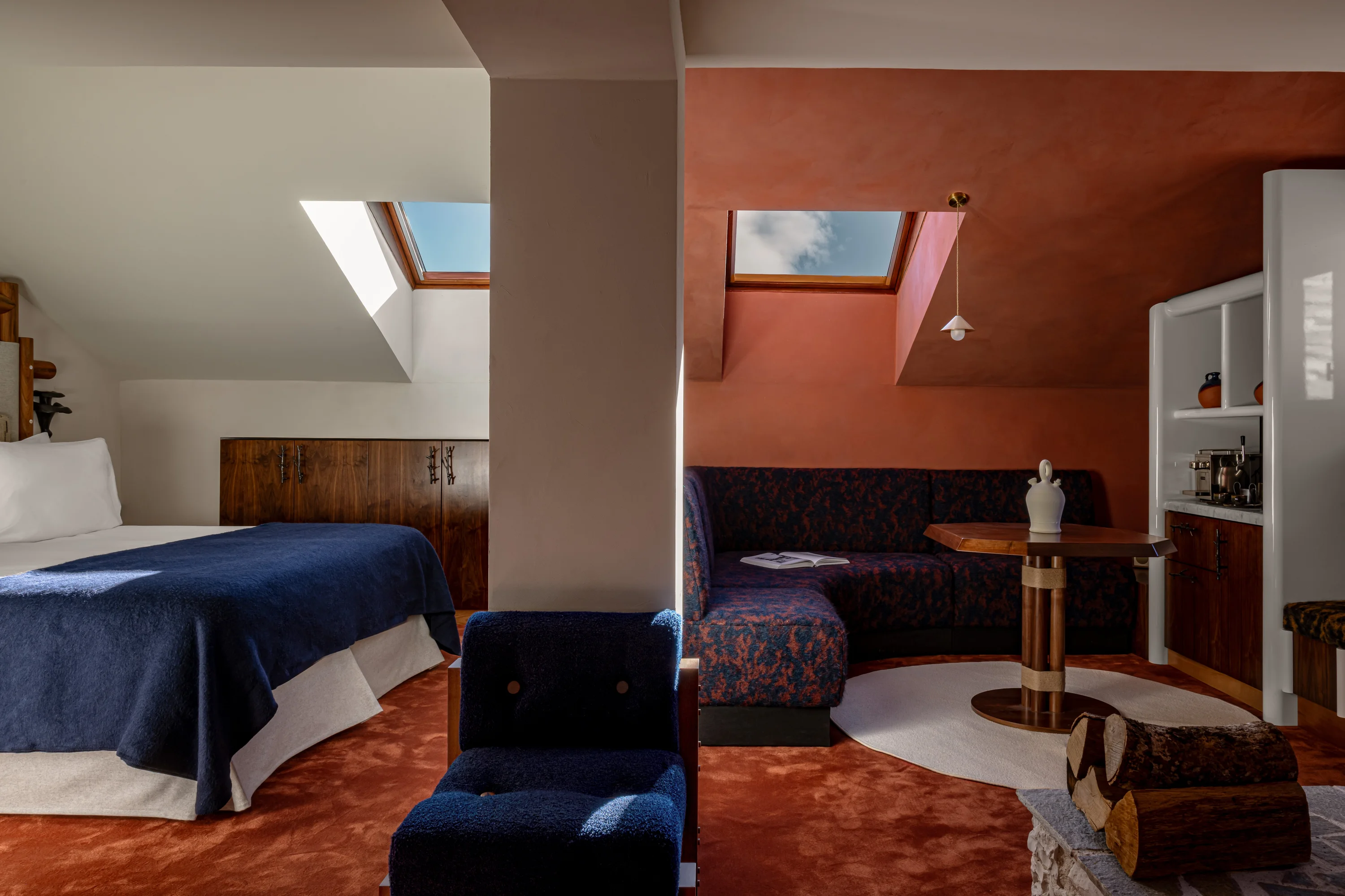 Hotel room with blue bedspread, navy chair, patterned sofa, wooden table, and skylight windows.