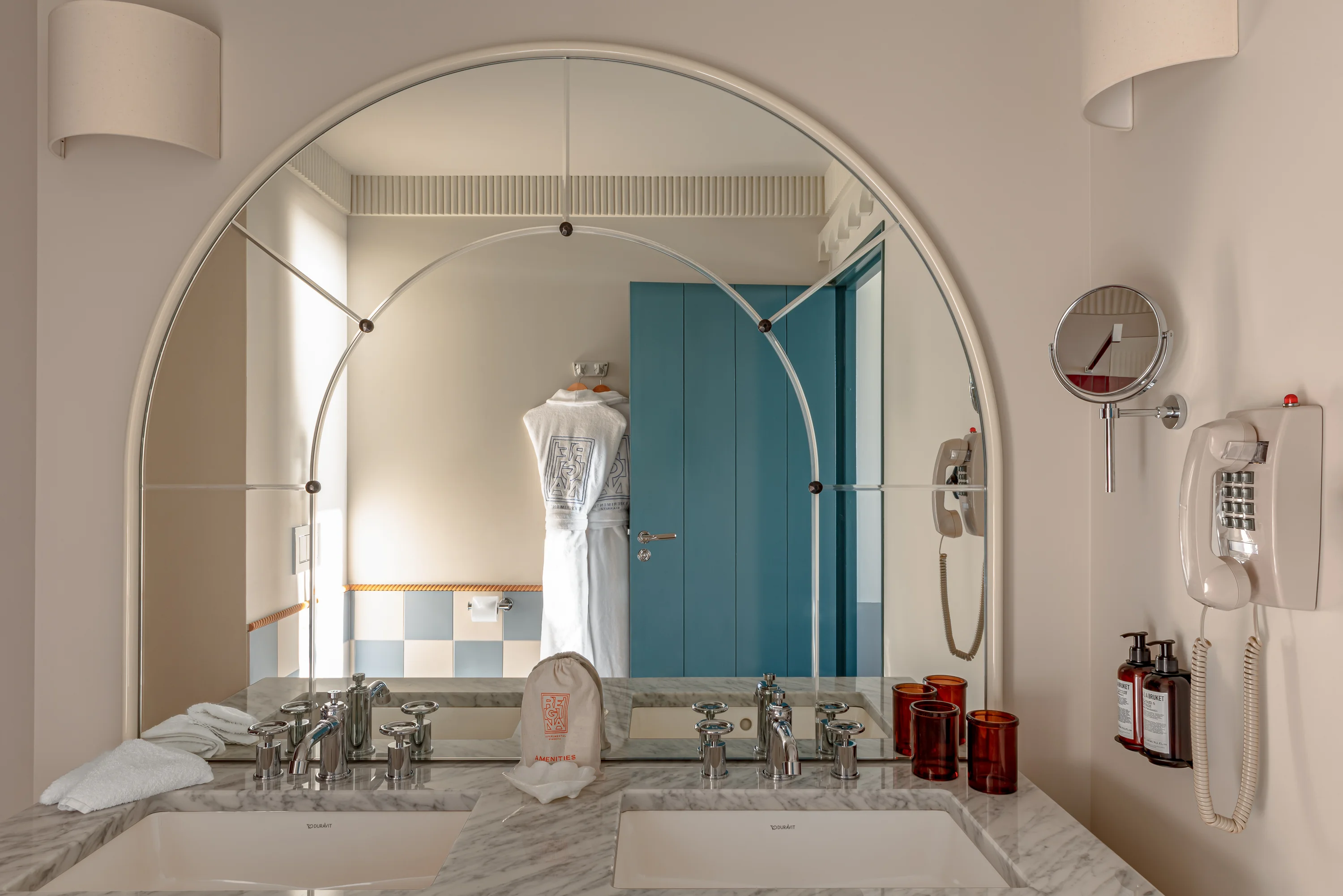 Bathroom with marble double sink, large arched mirror, blue door, hanging robe, and wall phone with toiletries.