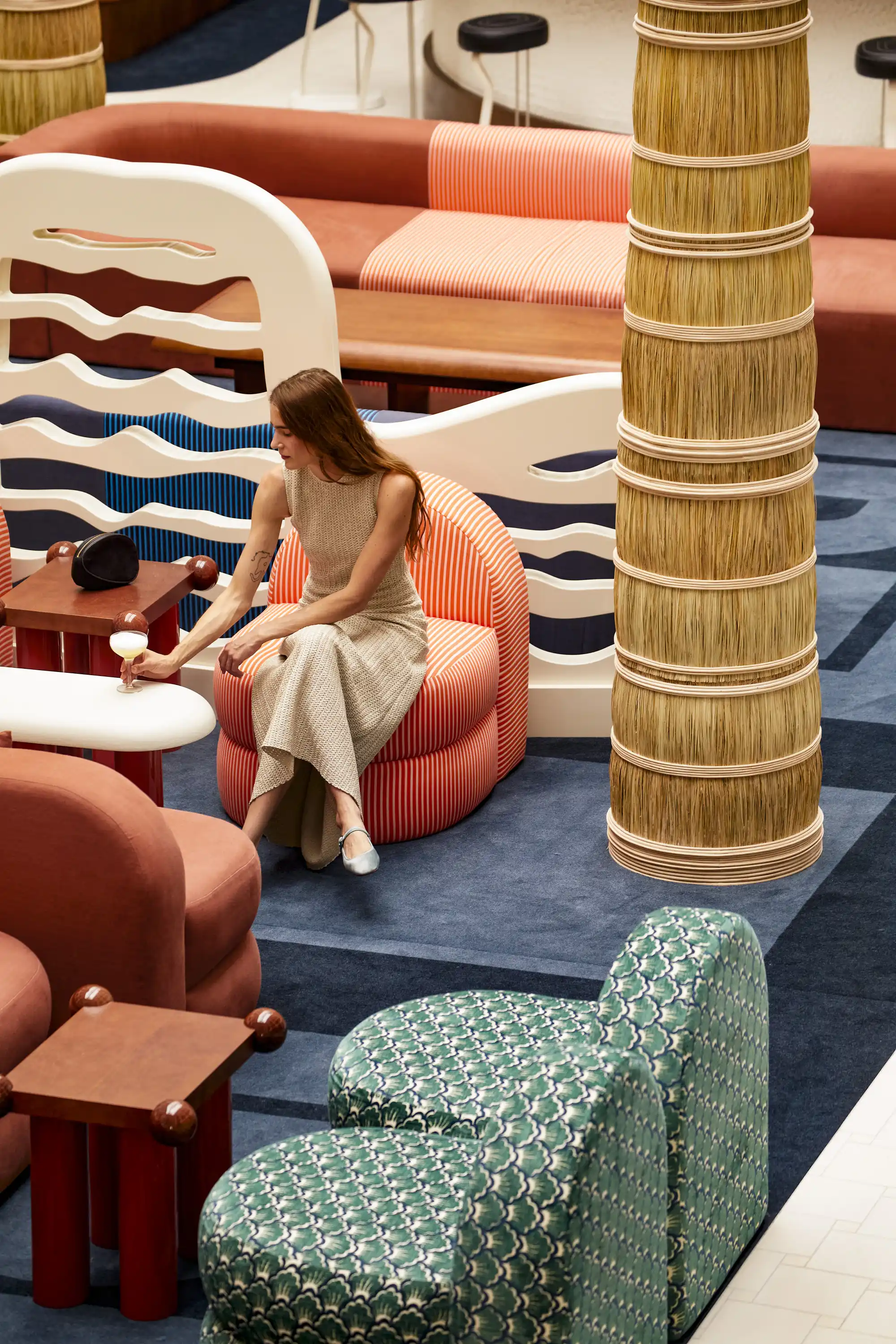 Woman in beige dress sitting on striped chair holding a cocktail in a colorful modern lounge.