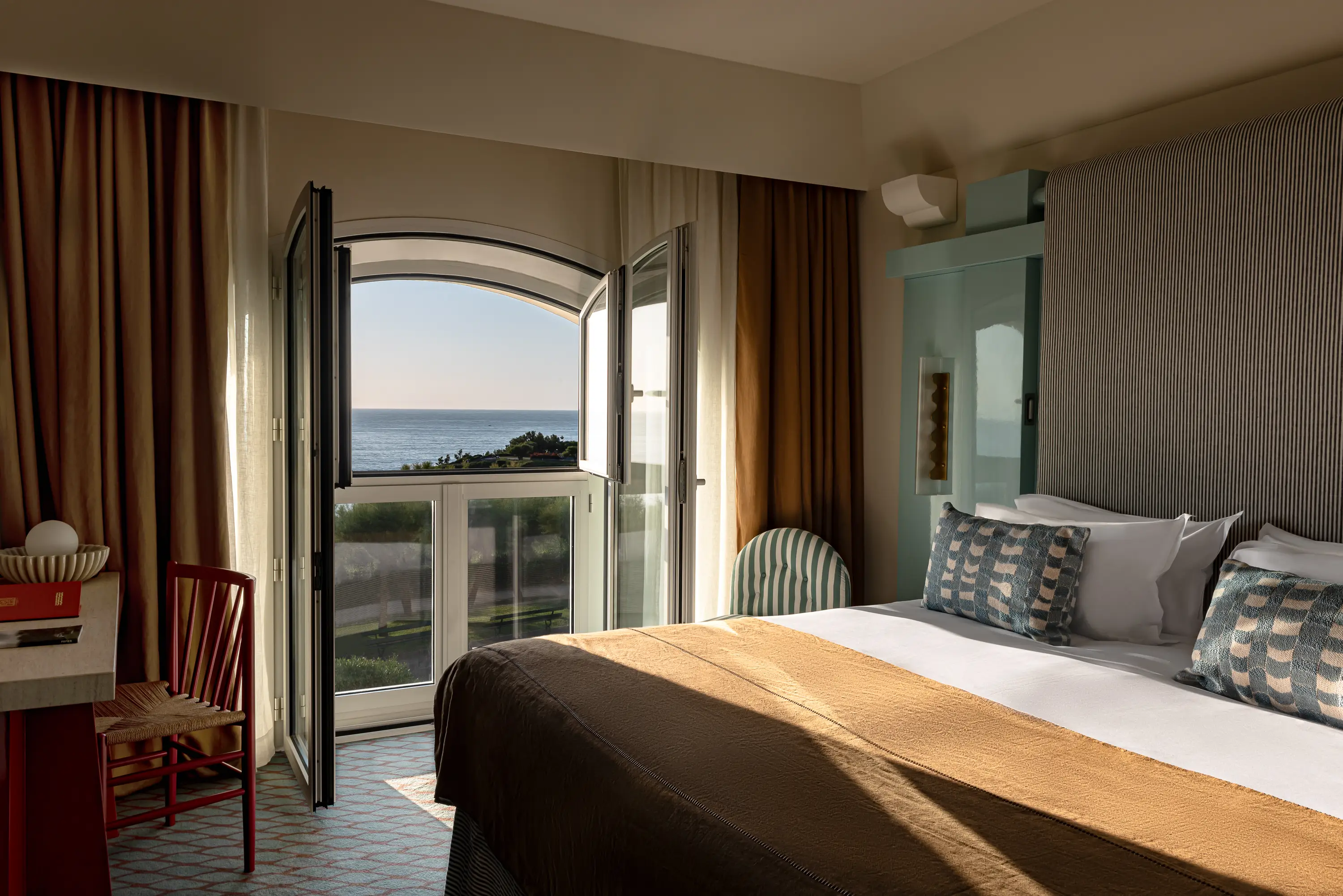 Hotel bedroom with double bed, striped pillows, open balcony door, and ocean view in sunlight.