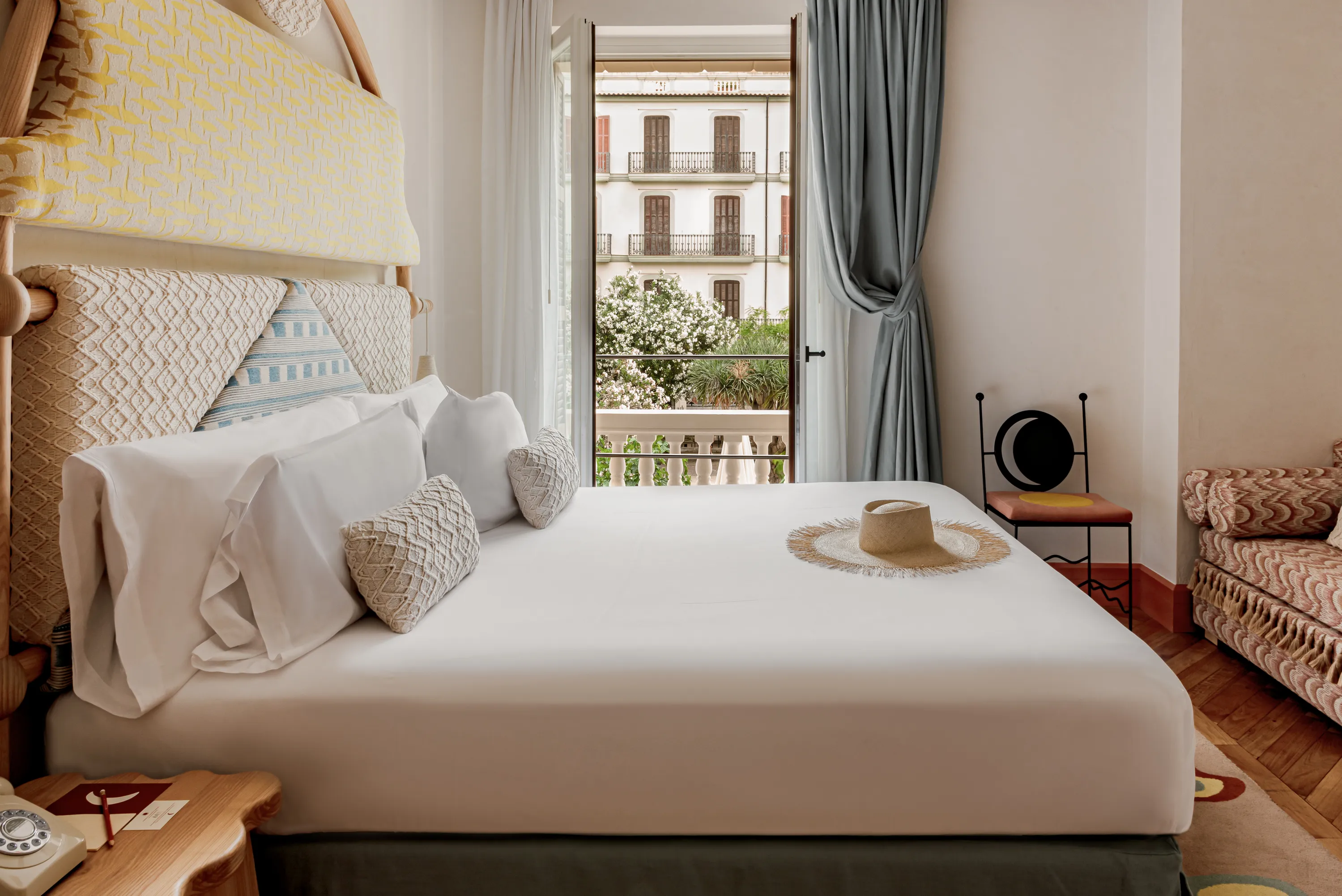 Bed with white linens, woven pillows, and a straw hat near an open balcony with greenery view.