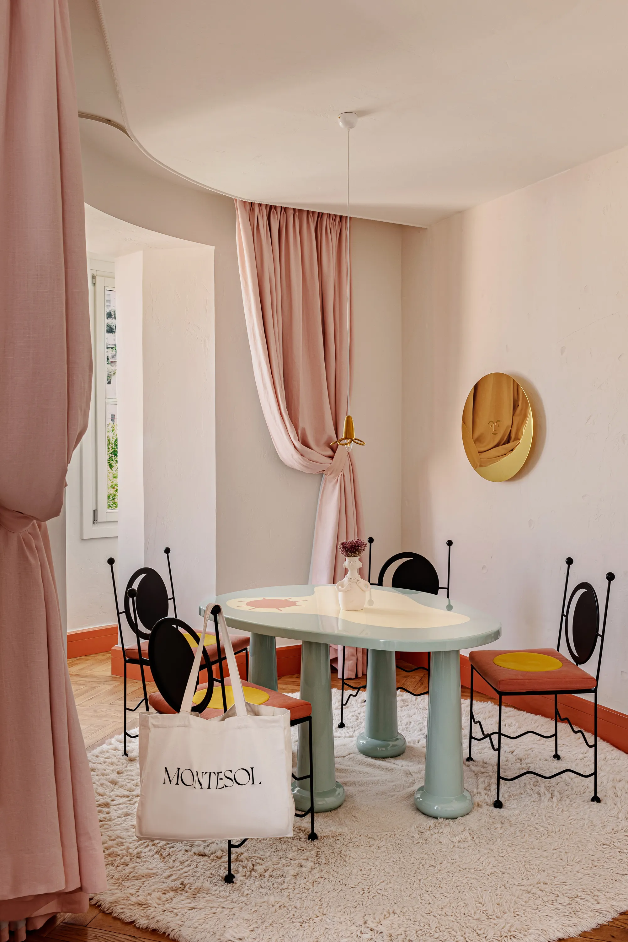 Dining room with round table, four chairs with yellow seats, pink curtains, and a bag labeled Montesol.