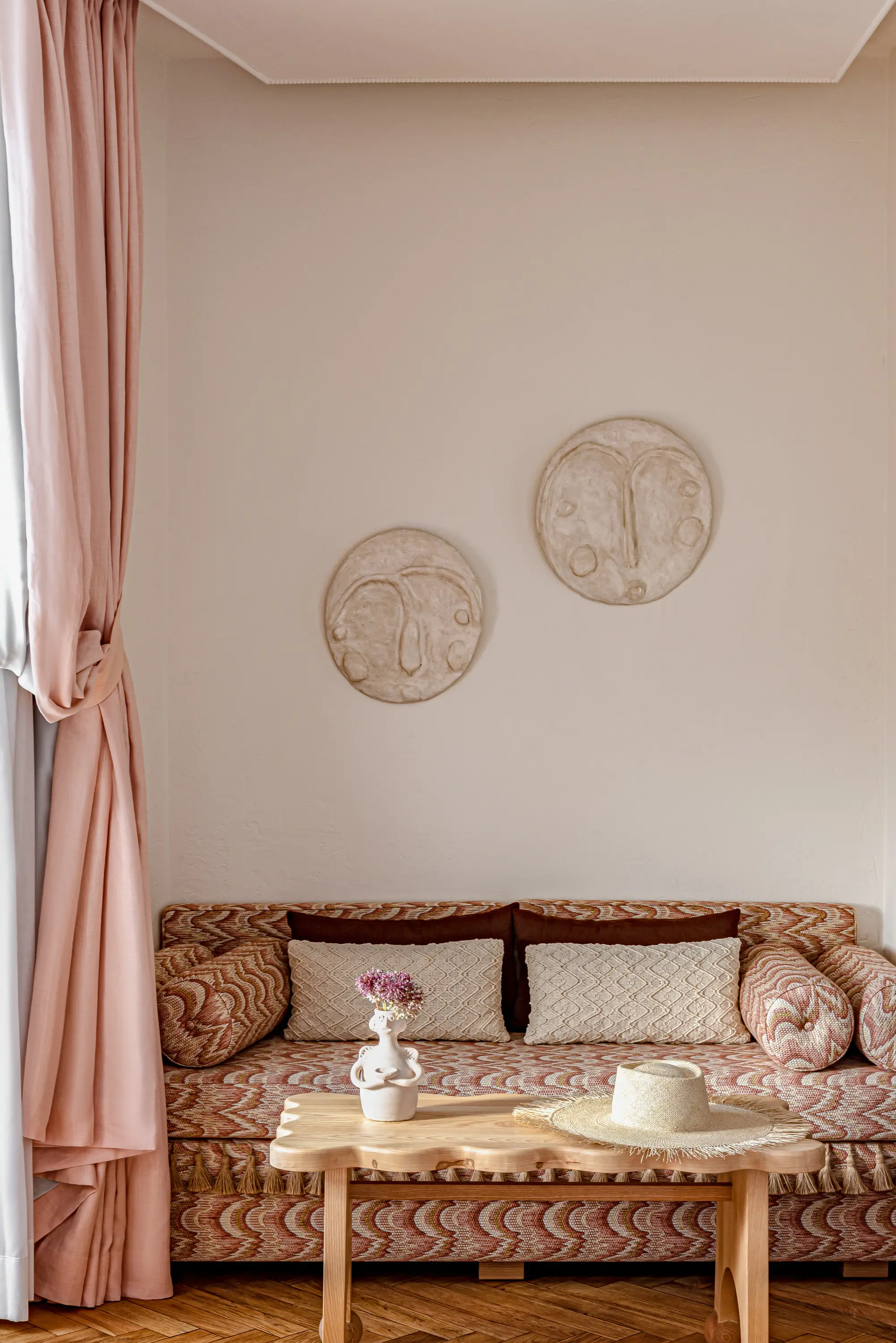 Textured patterned sofa with pillows, wooden coffee table, vase with flowers, straw hat, and pink curtains.