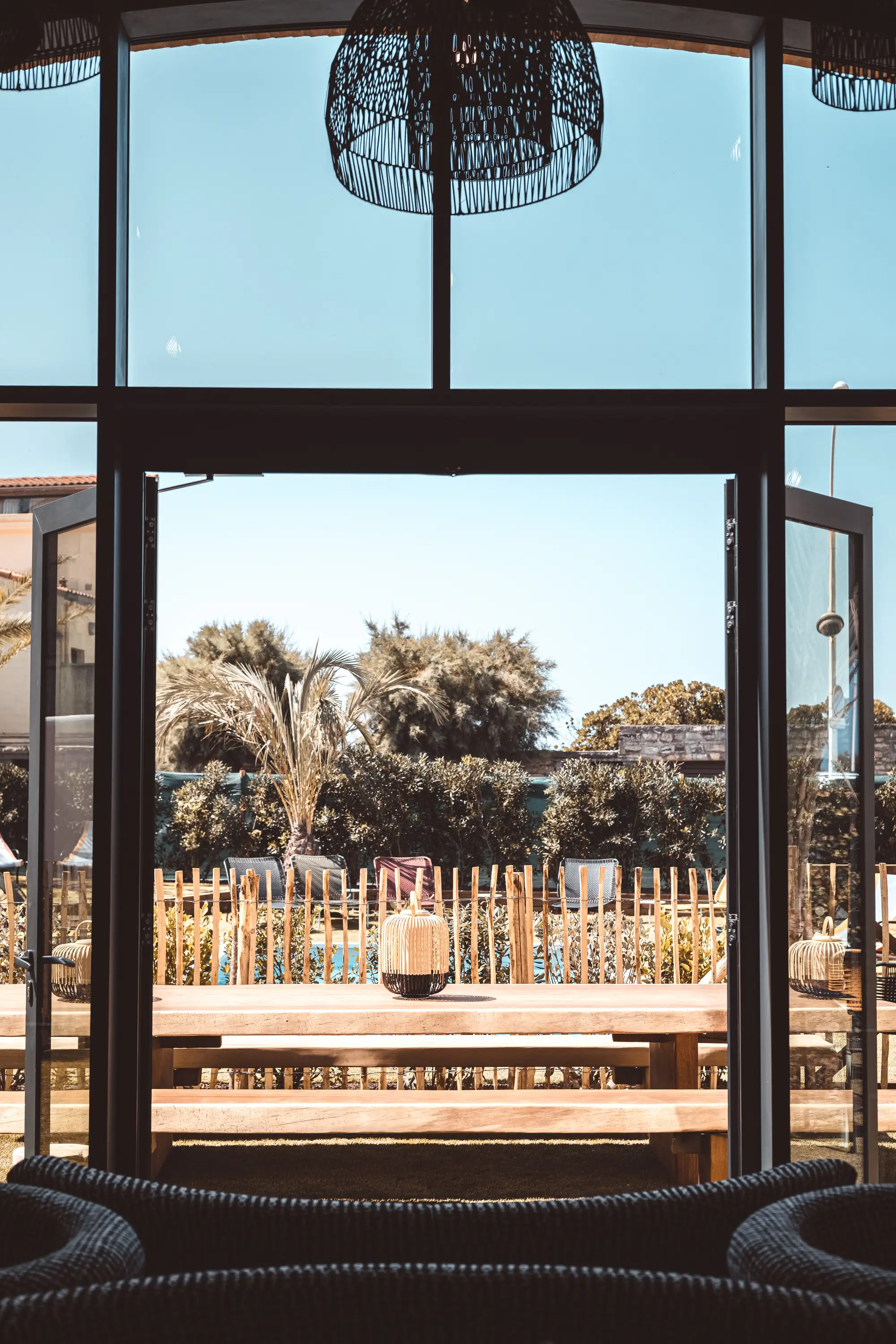 Wooden table and benches outside with wicker lanterns, viewed through open glass doors.