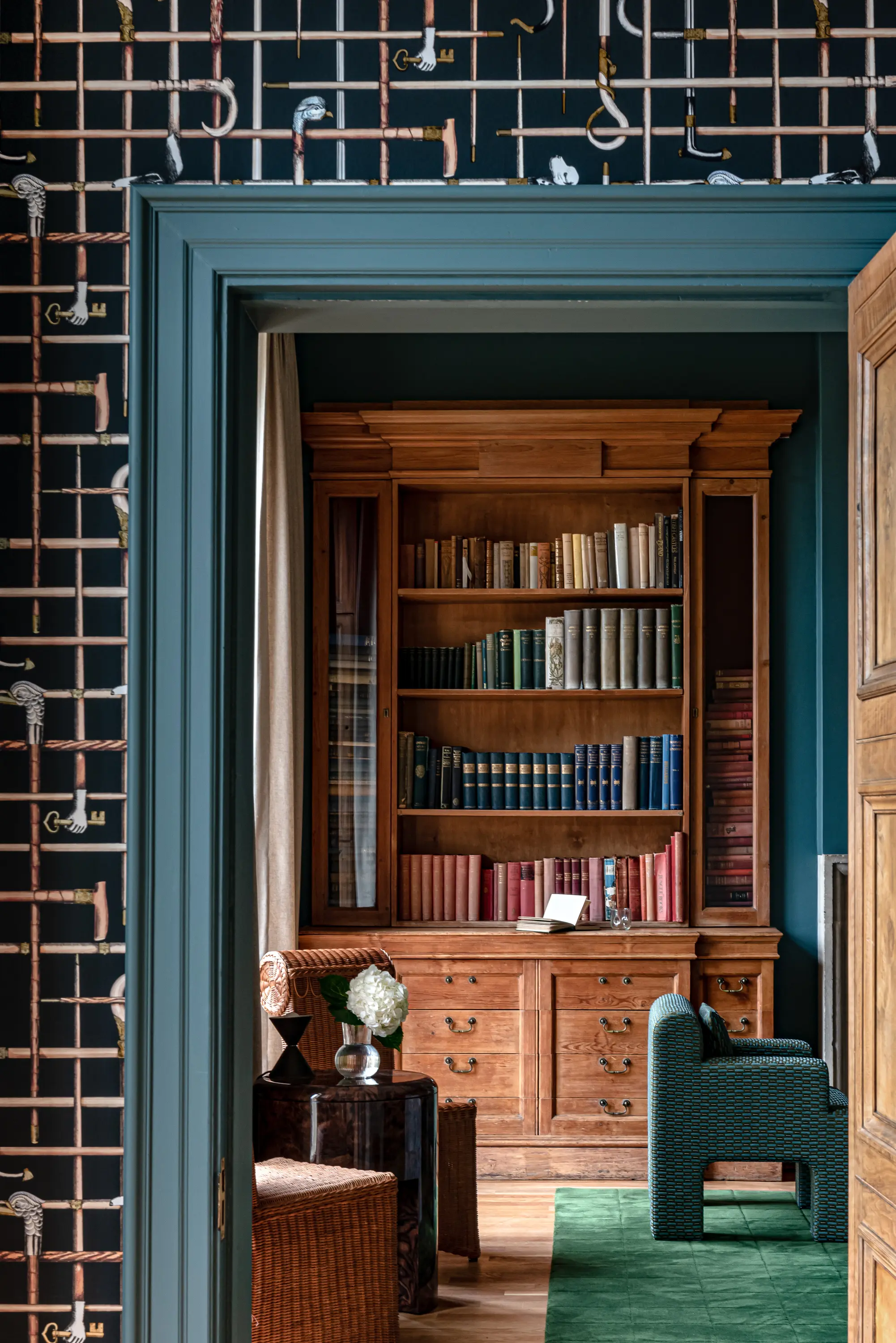 Wooden bookshelf with colorful books in a cozy room with patterned wallpaper and comfortable chairs.