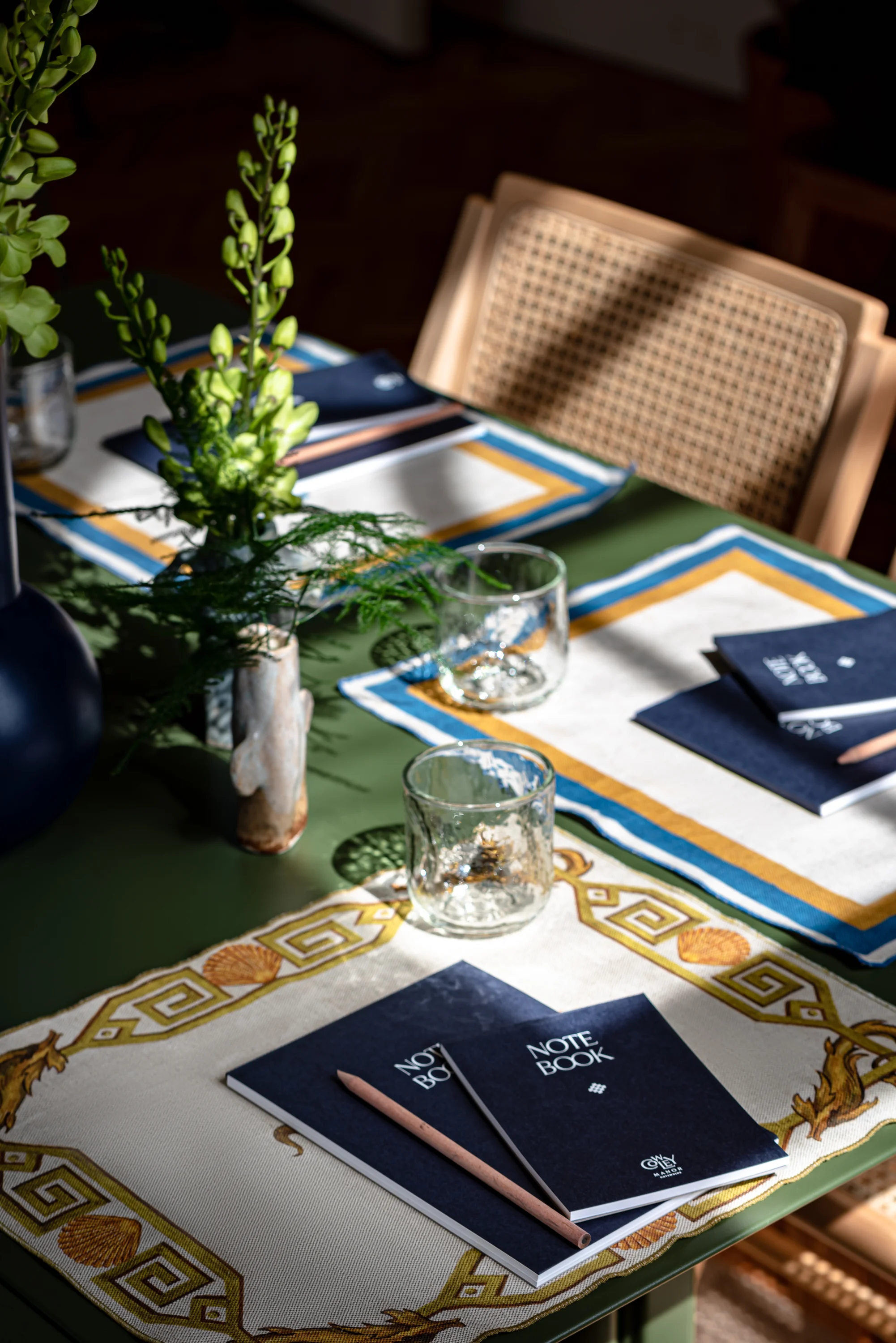 Restaurant table with decorative placemats, glass tumblers, green plants, navy notebooks, and wooden pencils.