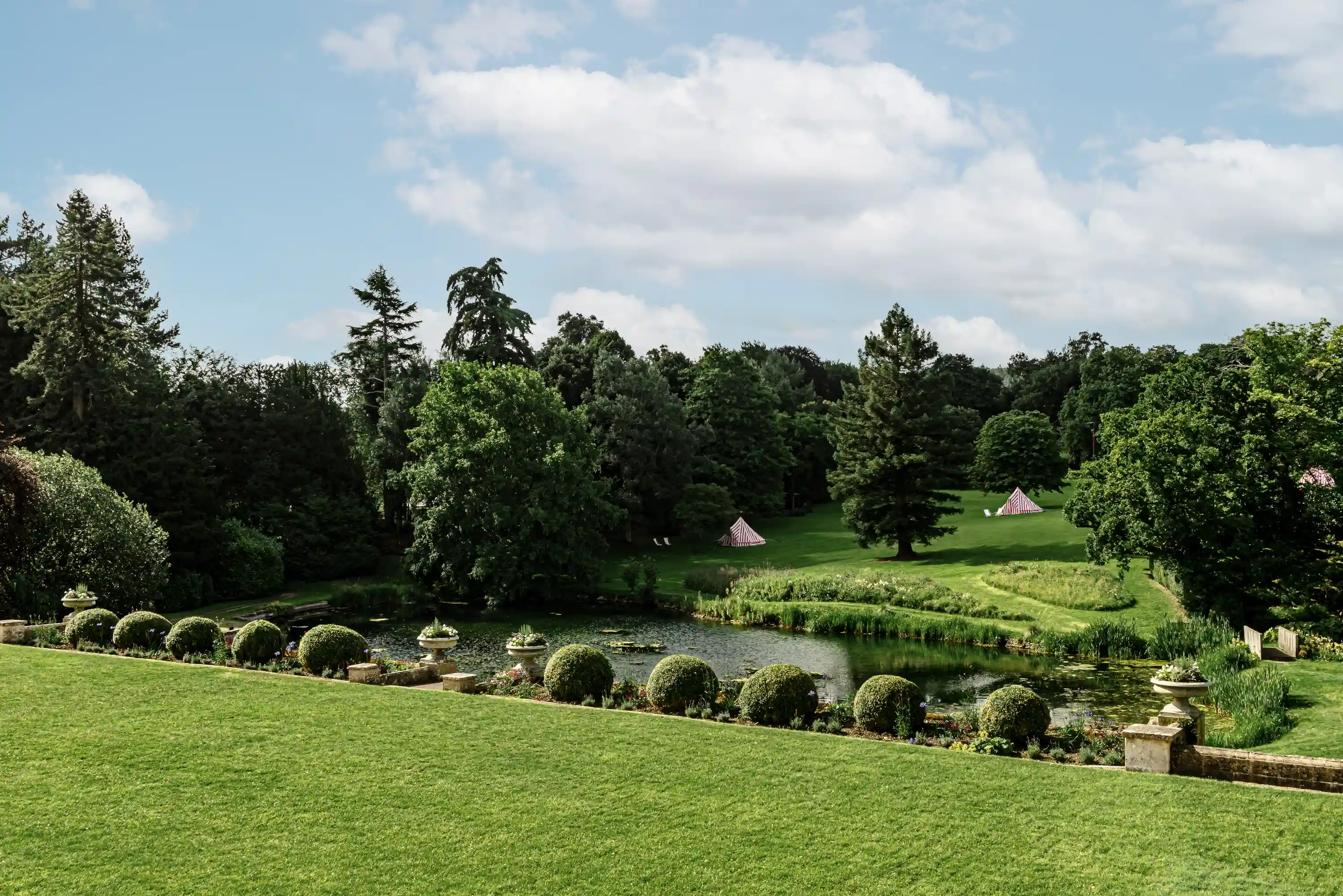 Green lawn with pond, manicured shrubs, trees, and red-and-white striped tents in the distance under a blue sky.